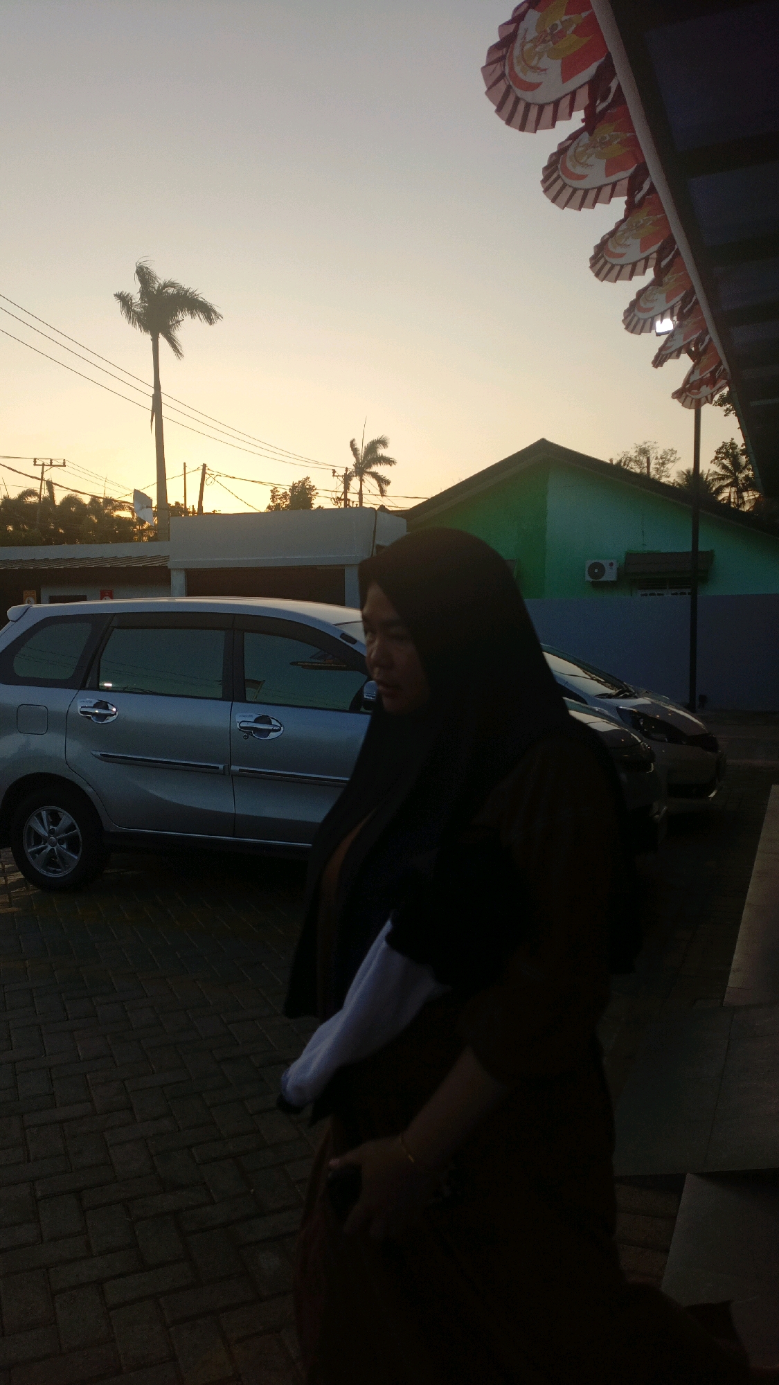 The image captures a side profile of a woman wearing a black hijab and a brown garment, standing on a paved walkway. She is positioned in the foreground, with her body angled slightly away from the camera. Behind her, a silver minivan is parked, and beyond that, a light green building with an air conditioning unit is visible. The background features a clear sky with a warm, gradient sunset. Several power lines and palm trees are also present, suggesting an outdoor urban or suburban setting. In the upper right portion of the image, a decorative banner with intricate patterns is partially visible, hanging from a structure. The overall lighting indicates it is late afternoon or early evening.
