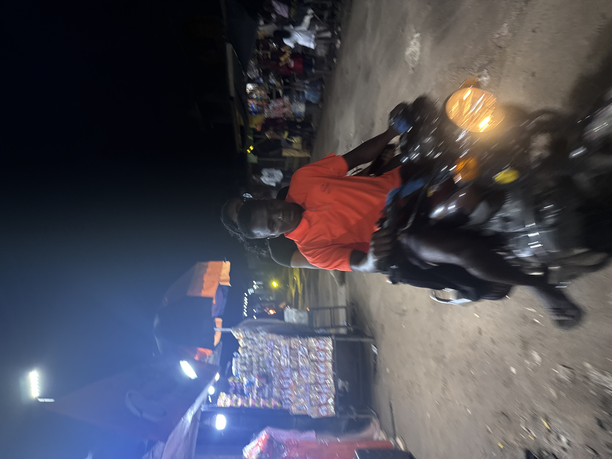 A low-angle, motion-blurred photograph captures two individuals on a motorcycle at night, likely in Ihiagwa, Nigeria. The person in the foreground, a young man wearing an orange polo shirt, is driving the motorcycle. He has short dark hair and is looking forward. Behind him, a person with braided hair, wearing a dark shirt, is a passenger.
The motorcycle has a bright, illuminated headlight. The setting appears to be a street or market area with various stalls visible in the background. On the left, a stall is packed with goods, possibly drinks or packaged food items, illuminated by a bright light. Umbrellas are set up over some of the stalls. The ground is dirt or gravel. People are gathered in the background, suggesting a community gathering or market activity. The overall atmosphere is active and somewhat crowded.