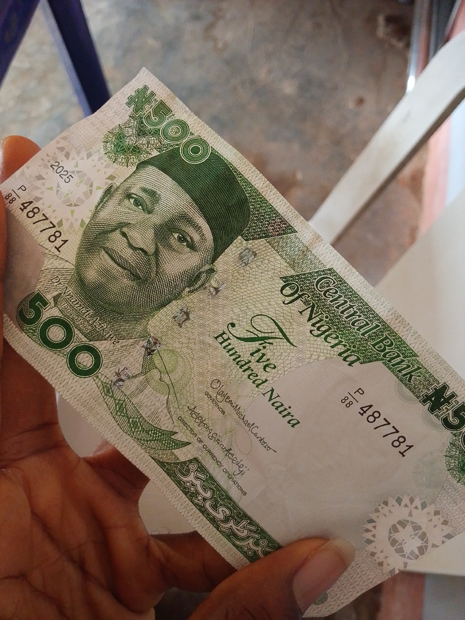 A hand holds a Nigerian 500 Naira banknote. The banknote features a portrait of Nnamdi Azikiwe. The denomination "500" is prominently displayed, along with the text "Central Bank of Nigeria" and "Five Hundred Naira." Serial numbers are visible, including "P 88 487781". The background appears to be an indoor setting, possibly a room with a textured floor and furniture, though no specific landmarks of Lagos are discernible. The lighting suggests it is daytime.