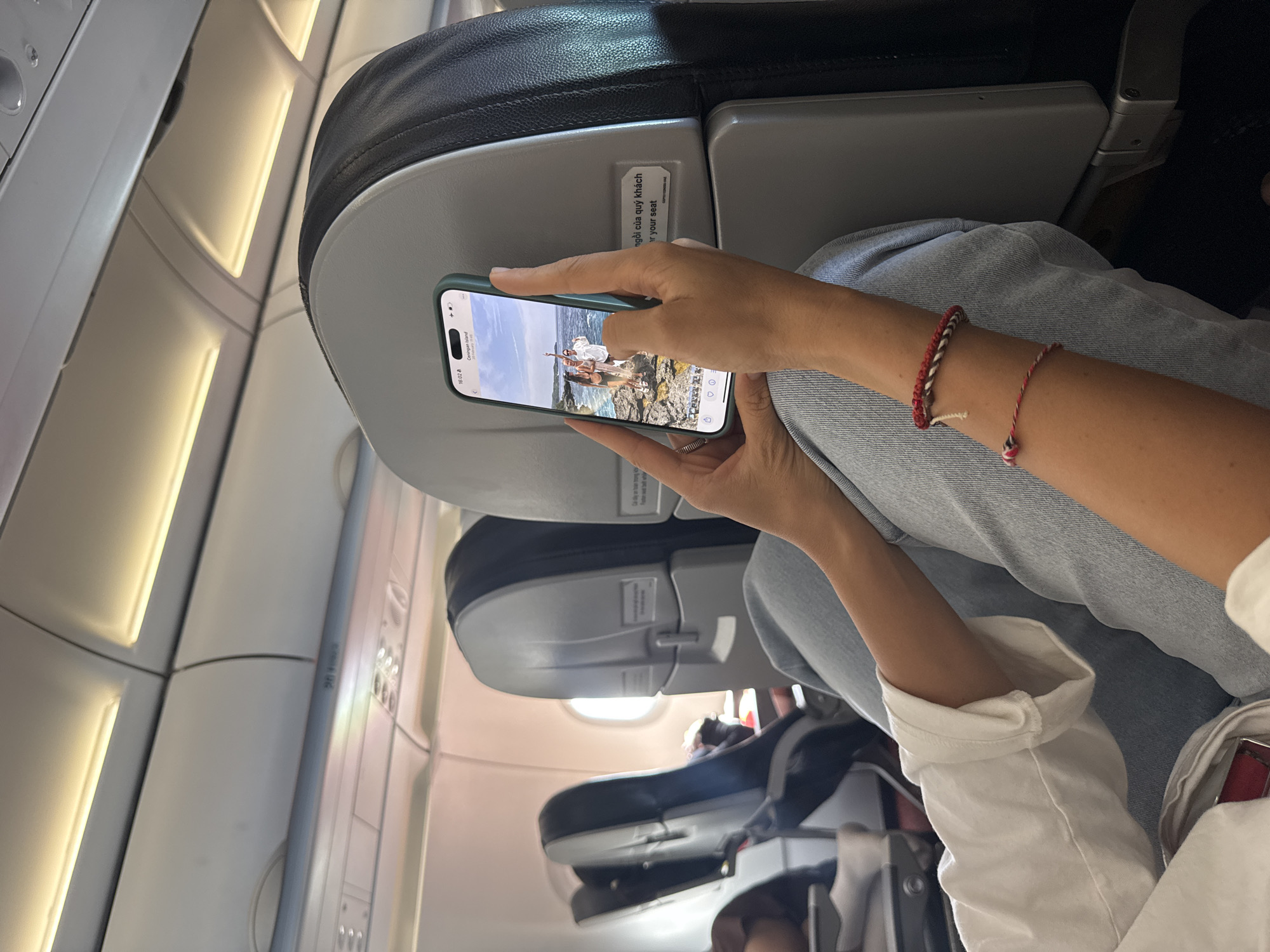 The image depicts the interior of a commercial aircraft cabin, indicating a setting at or around Đà Nẵng International Airport (DAD) in Đà Nẵng, Vietnam. In the foreground, a passenger is shown from a seated position, wearing a white long-sleeved shirt with rolled sleeves and light blue jeans. Two red and white braided string bracelets are visible on their right wrist. The passenger holds an iPhone, identifiable by its Dynamic Island feature and a light green protective case, with both hands. The phone screen displays a full-screen photograph of three individuals – two women in swimwear and a man in a white shirt – standing on a rocky coastal cliff overlooking the ocean under a clear sky. The photograph is geotagged as "Ceningan Island" and dated "21 February 2023." The phone's status bar indicates the time as 16:02. The cabin environment includes grey and black airplane seats, overhead compartments, and individual reading lights. A window showing bright exterior light is visible in the background. A sticker on the seatback directly in front features text in Vietnamese, "ngồi của quý khách," followed by its English translation, "your seat." A partially visible passenger occupies a seat further to the left.