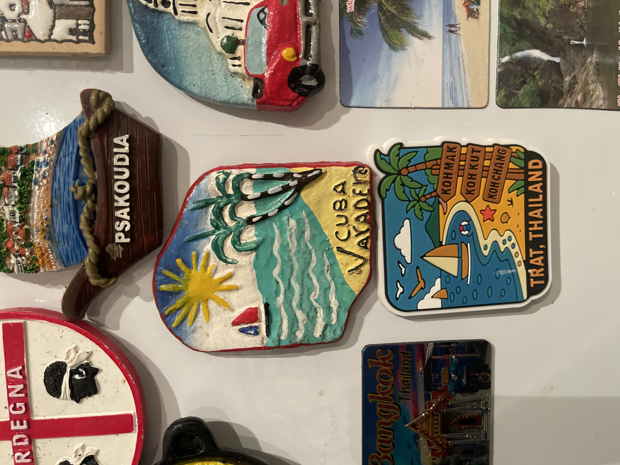 The image shows a collection of souvenir magnets attached to a white surface, possibly a refrigerator. The magnets represent various locations, including Sardegna (Sardinia, Italy), Psakoudia, Cuba (Varadero), Bangkok (Thailand), and Trat (Thailand), specifically Koh Mak, Koh Kut, and Koh Chang. The magnets vary in shape, size, and material, with some being ceramic, plastic, or photographic prints. There are no people present in the image, only inanimate objects. The location is not in Maidstone, United Kingdom.