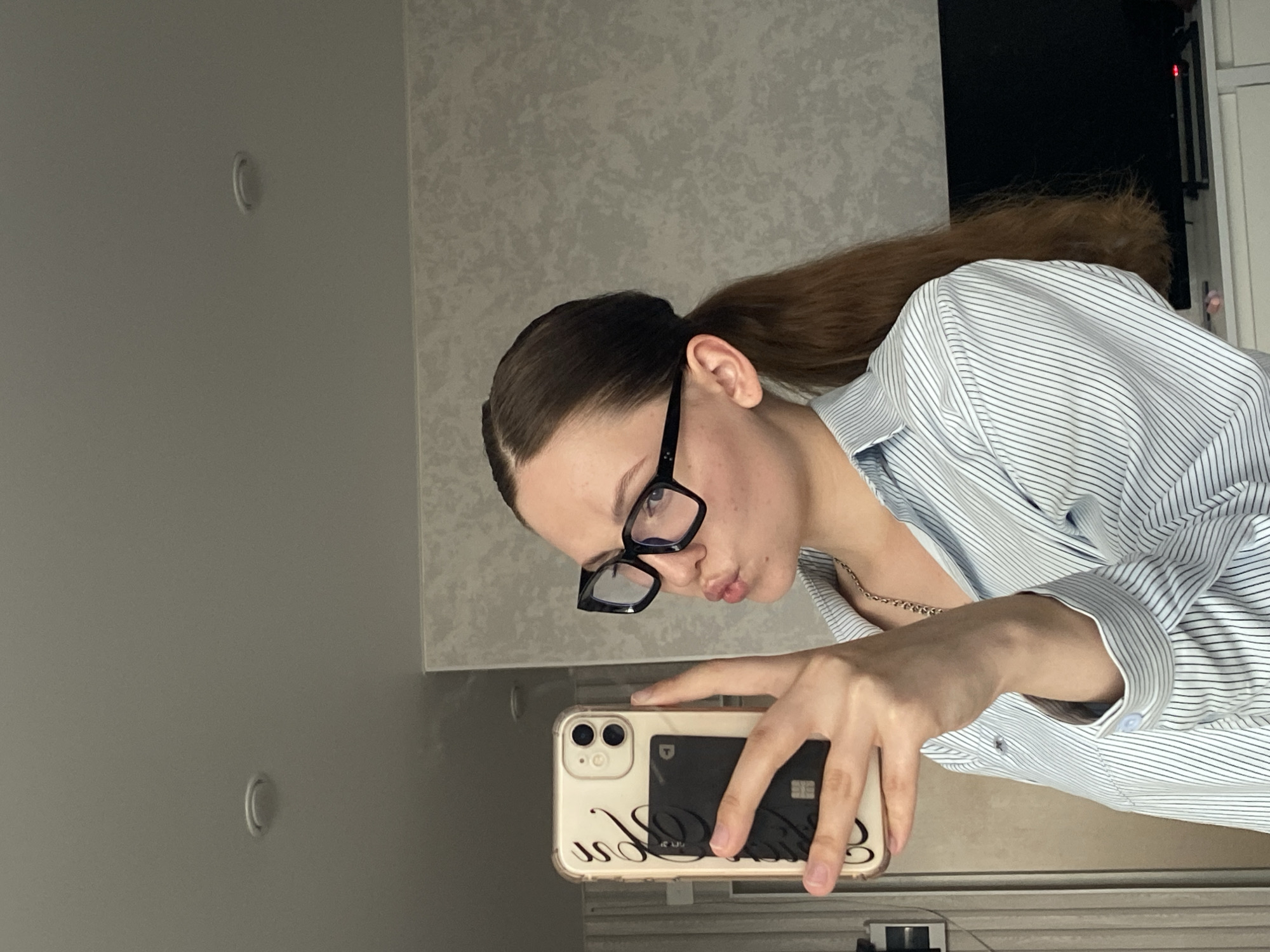 A young woman with dark brown hair in a ponytail, wearing black-rimmed glasses and a blue and white striped button-down shirt, takes a selfie in a dimly lit room. She is holding a white smartphone with a clear case, which has a black card holder attached to the back. The woman is puckering her lips as she looks into the phone's camera. The background appears to be a residential interior, with a textured grey wall and a ceiling light visible. Based on the provided context, the location is Izhevsk, Russia.