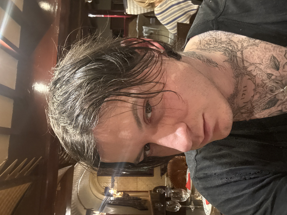 The image displays a close-up indoor portrait of a male individual, appearing to be in their 20s or 30s, situated within a restaurant or dining establishment in Deal, United Kingdom. The subject has dark, medium-length hair, which appears disheveled. They possess light-colored eyes and maintain direct eye contact with the viewer, exhibiting a neutral to serious facial expression. Extensive black ink tattoos cover the individual's neck and upper chest, featuring detailed patterns and the number "1711" on the right side of the neck. A thin silver chain with a circular pendant hangs around their neck. They are wearing a dark, possibly black, short-sleeved top with visible distressed areas on the left shoulder.
The background reveals a warmly lit interior. To the immediate left, a table is set with two empty stemmed wine glasses, one water glass, a white plate with a blue rim, and a folded red napkin. A dark brown wicker chair back is partially visible behind these items. Further into the left background, an ornate arched alcove with a patterned wall covering and a wall-mounted sconce is visible. The ceiling features dark wooden beams. In the far right background, another individual, out of focus, is seated at a table, identifiable by a horizontally striped garment covering their back and shoulder. No direct interaction between individuals is depicted.