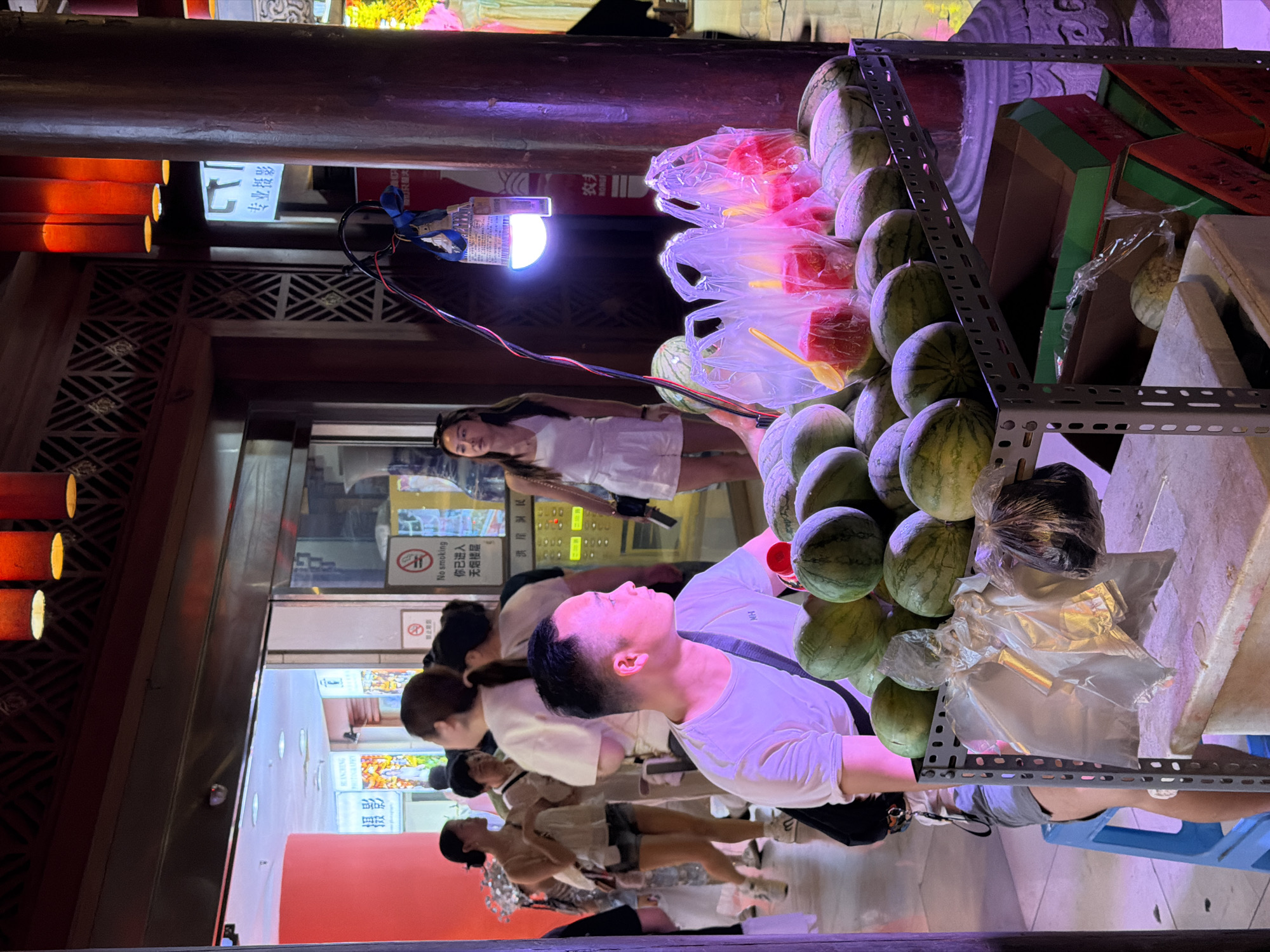 The image depicts a bustling street scene at night in the Yuzhong District of China. In the foreground, a man in a white t-shirt sits on a blue stool behind a cart filled with watermelons. He appears to be a vendor, looking off to the side. The cart also holds several plastic bags containing cut watermelon pieces, each with a yellow plastic spoon. In the background, several people are walking past or standing around, engaged in various activities. One woman in a white tank top and shorts is visible on the left, while another woman in a white dress stands further back. The environment is characterized by traditional Chinese architecture, with wooden pillars and decorative elements, illuminated by the warm glow of overhead lanterns and a bright LED light above the watermelon cart. Signage in Chinese characters indicates the presence of shops and services, including one that appears to be a photo studio. The overall atmosphere suggests a vibrant commercial area with a mix of locals and possibly tourists.