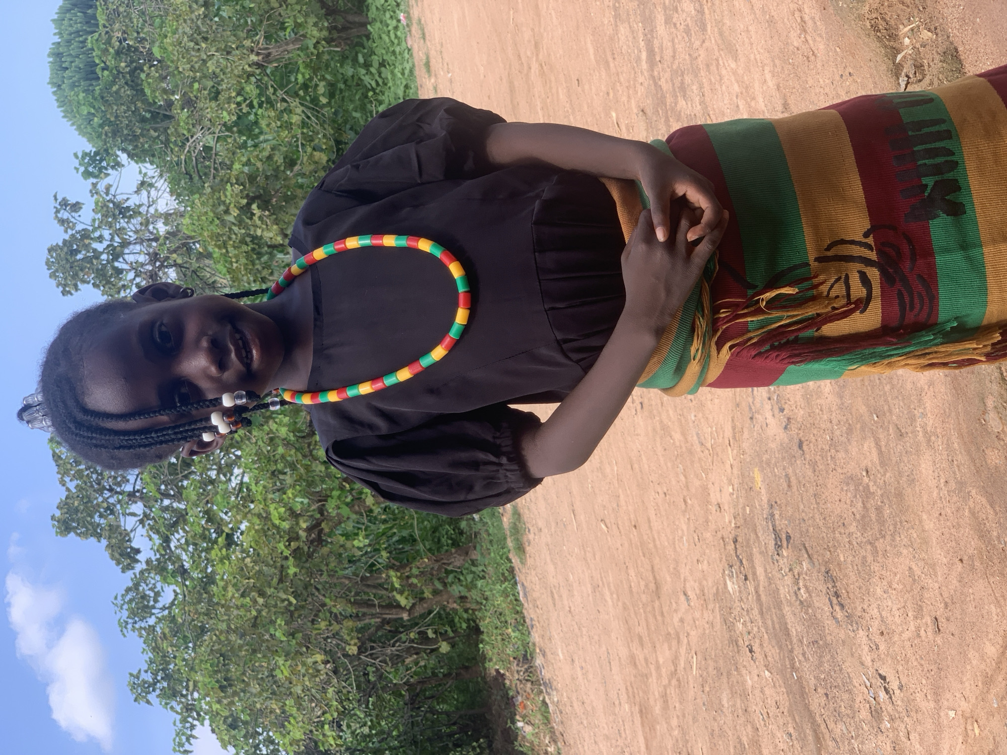A young girl with braided hair adorned with beads and a colorful beaded necklace stands in front of lush green trees under a cloudy blue sky. She is wearing a black puffed-sleeve top and a vibrant, striped skirt with red, green, and yellow bands. The skirt features fringed accents and a black graphic design. Her hands are clasped in front of her. The ground is a reddish-brown dirt path, suggesting an outdoor environment in Sabon Tasha, Nigeria.