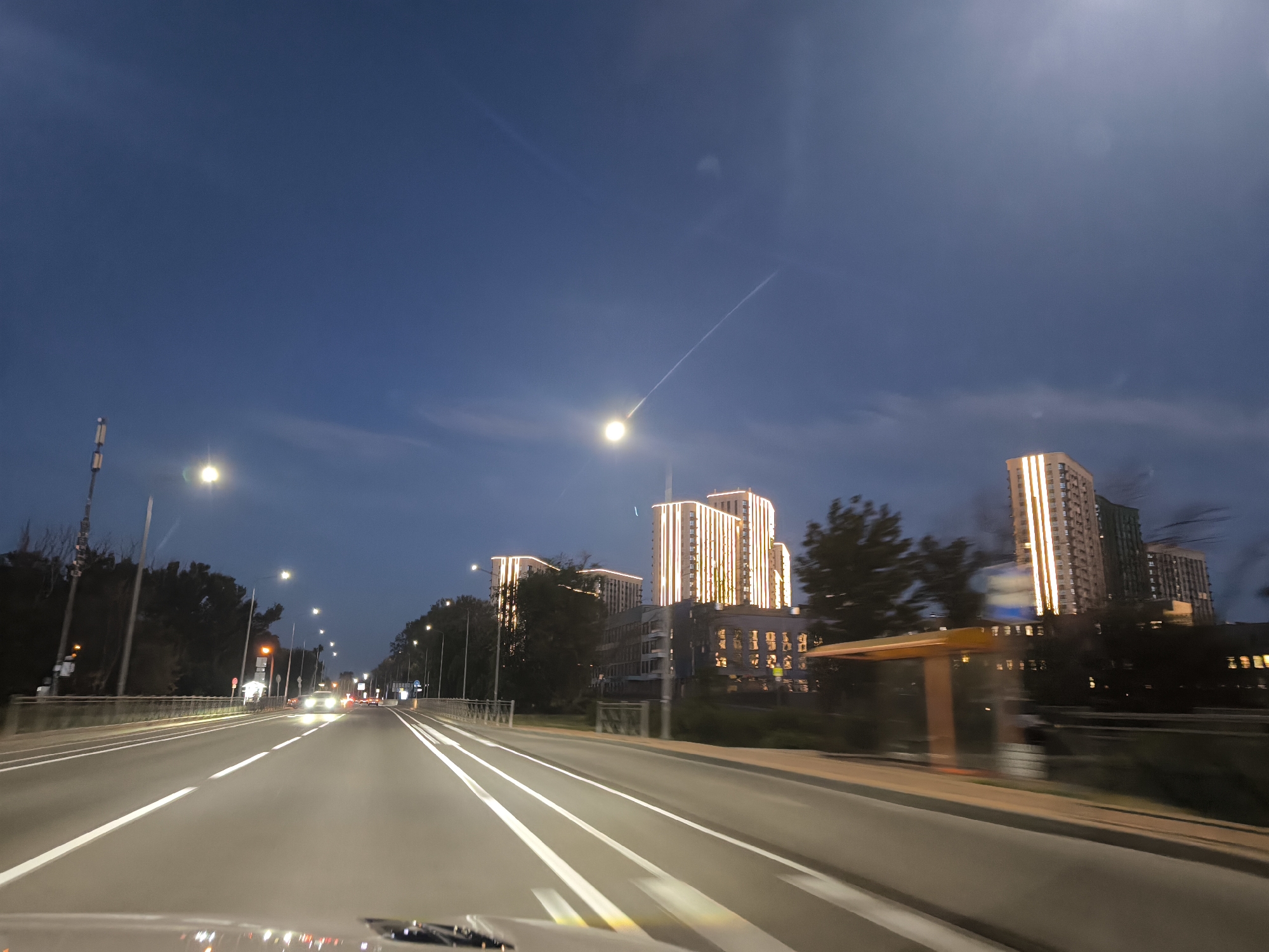 A wide-angle shot taken from the interior of a moving vehicle, looking forward along a multi-lane road at dusk. The road has white lane markings. Streetlights line the road, casting a glow. In the distance, several tall, modern buildings are illuminated with vertical strips of light. Trees and other foliage are visible along the sides of the road. The sky is a deep blue, with faint contrails and a bright light, possibly a streetlamp or distant aircraft, visible in the upper part of the frame. A bus stop shelter with a yellow roof is visible on the right side of the road. The overall impression is an urban scene at twilight, with a sense of motion due to the perspective of the vehicle.