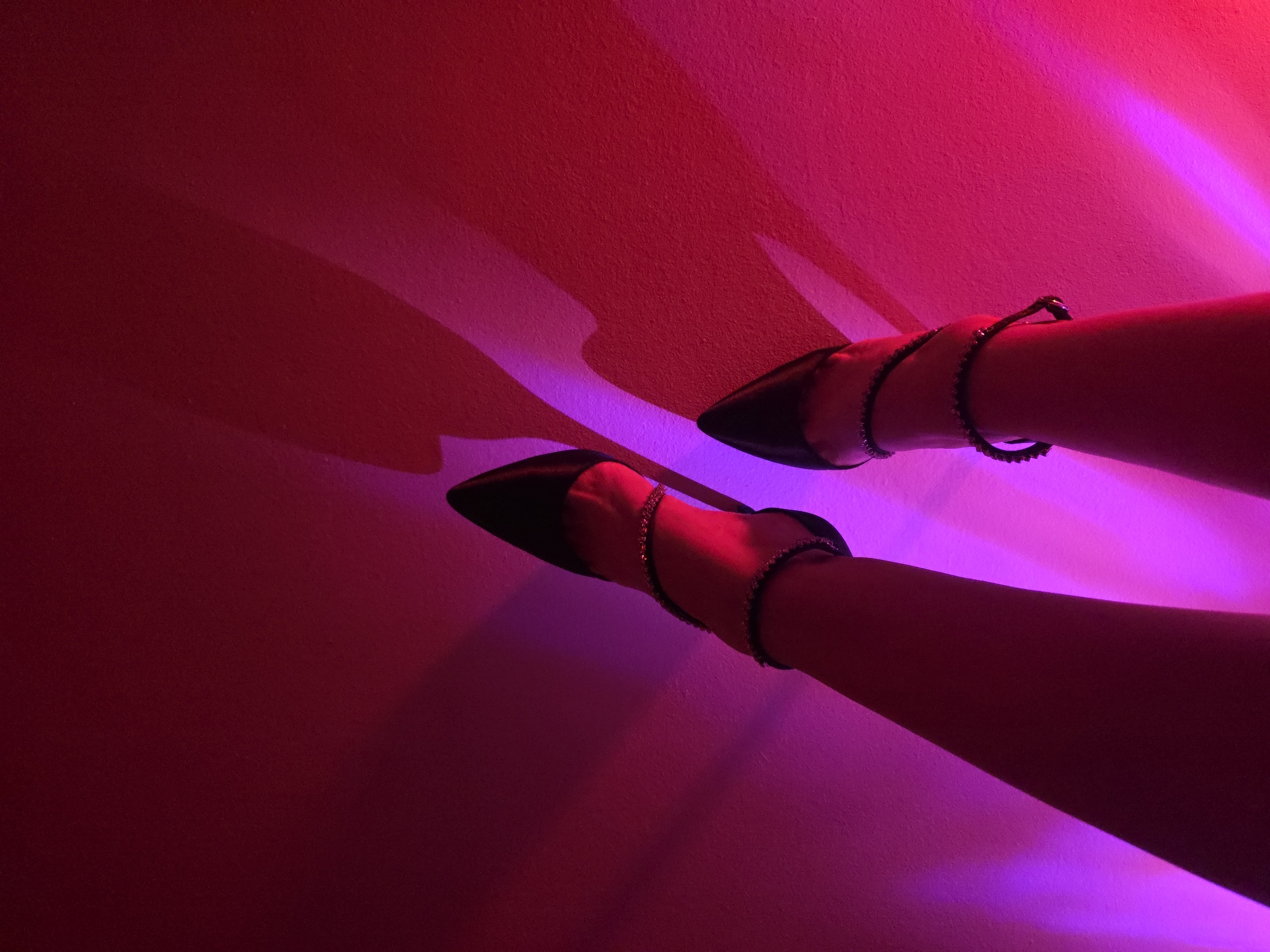 A high-angle shot depicts a pair of legs adorned with black pointed-toe heeled shoes and embellished ankle straps, set against a backdrop bathed in red and purple light. The specific location in the city of Unknown, Unknown, is not discernible from the provided image.