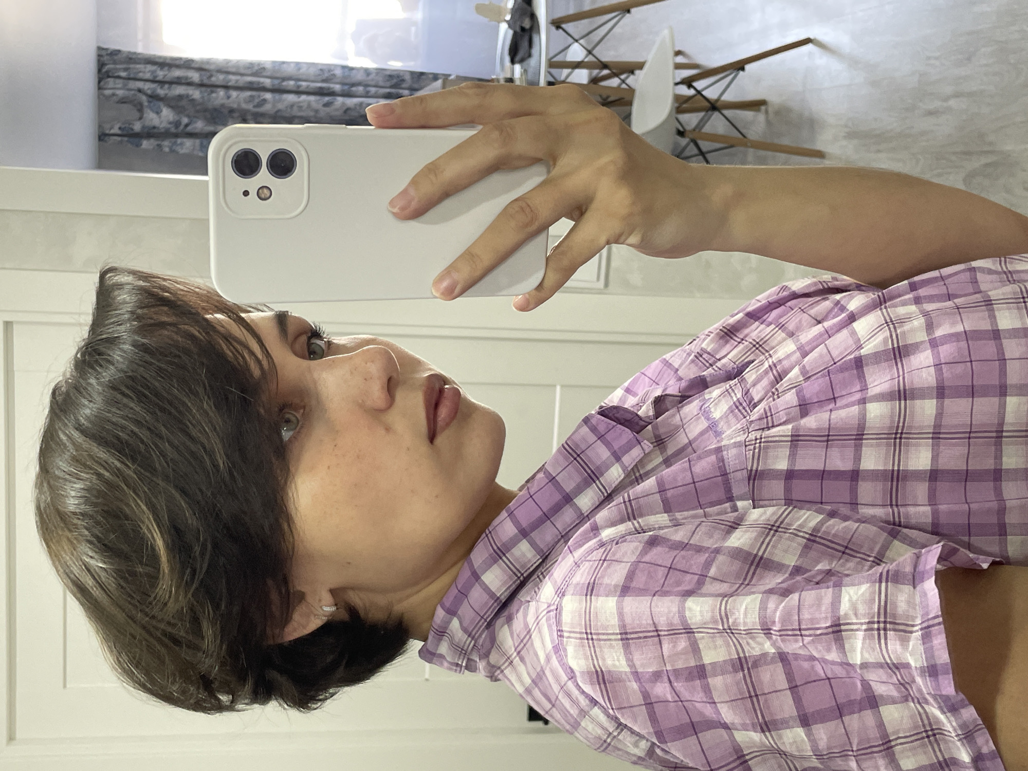 A woman is taking a selfie with a white smartphone. She is wearing a purple and white plaid shirt. She has short brown hair and light skin. The background includes a window with a curtain, a white table, and white chairs. The location appears to be an indoor space with modern furniture. There is no visible evidence to determine the city or country.