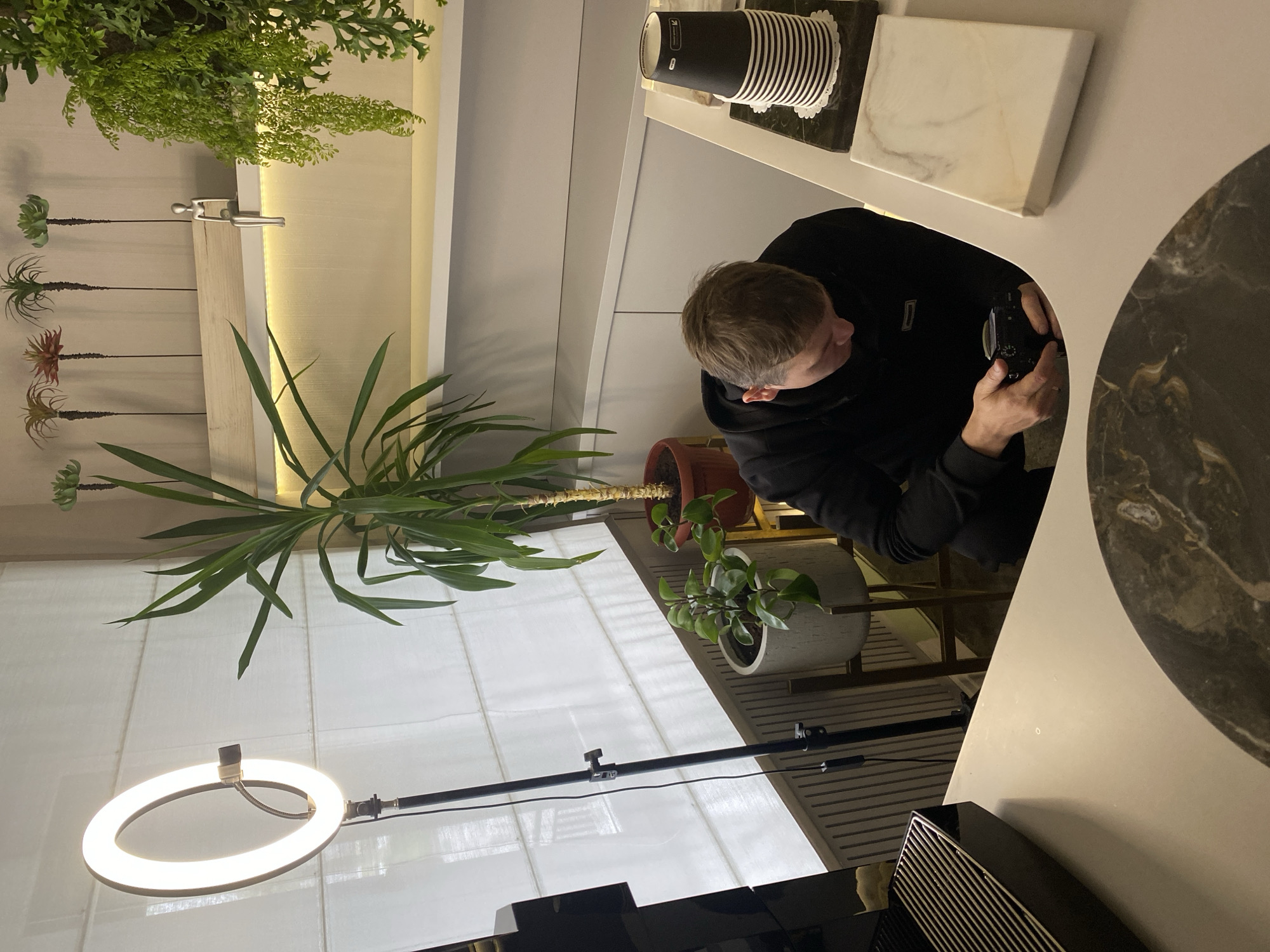 The image shows a room interior with various elements:
**Person:** A man in a black sweater is holding a camera, potentially taking a photograph.
**Objects:** There are several plants, including one large plant positioned prominently. A ring light stands on a black rod, which is positioned by a window covered by a white curtain. Additional objects include paper cups, a marble block, and a black object with a grill pattern, all set on a table.
**Environment:** The location appears to be indoors, possibly a cafe or studio setting, judging by the mix of plants, furniture, and lighting equipment. A window is covered with white curtains, providing diffused light.
**Location:** There is not enough information to verify whether this photo was taken in Votkinsk, Russia.