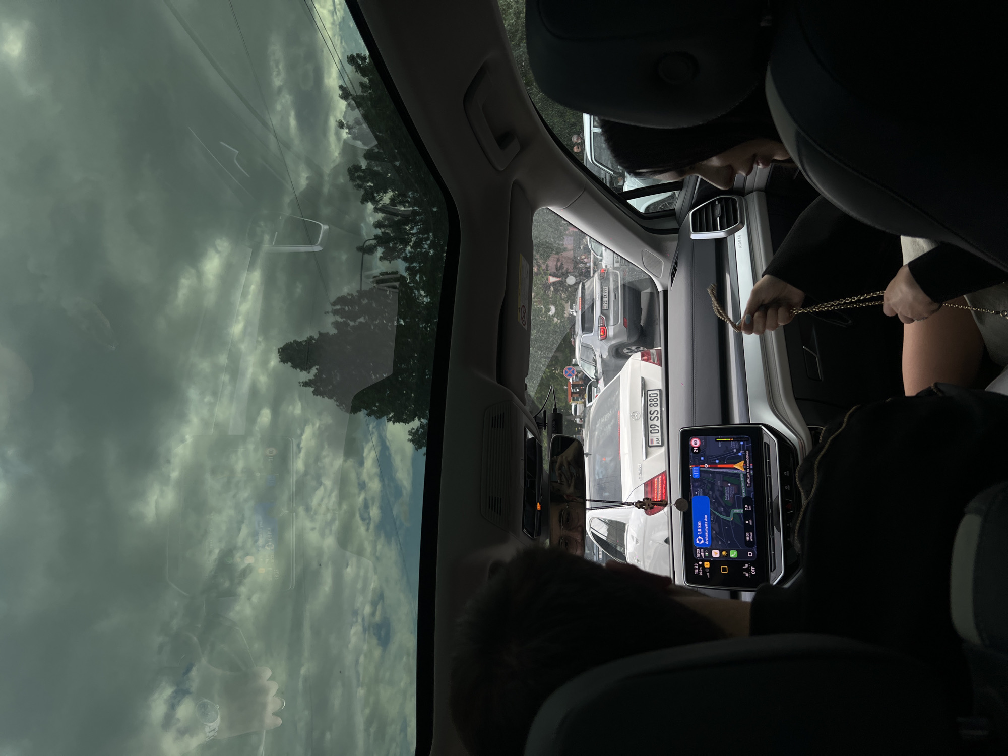 Here is a forensic description of the image:
The image is taken from the interior of a car, presumably from the back seat looking forward. The car is likely stopped in traffic, with other cars visible through the windshield. The license plate "09 SS 880" of a car in front is visible and identified as belonging to Armenia. The GPS/navigation screen on the dashboard indicates a location in Yerevan, Armenia, specifically noting "Arshakunyats Ave". The screen also indicates traffic, with a "Traffic jam for 4 min". The time displayed on the screen is 18:23.
A person with a gold chain is visible in the front passenger seat. An additional person is seated in the driver's seat. The car has a panoramic sunroof visible above. The sky is overcast.
