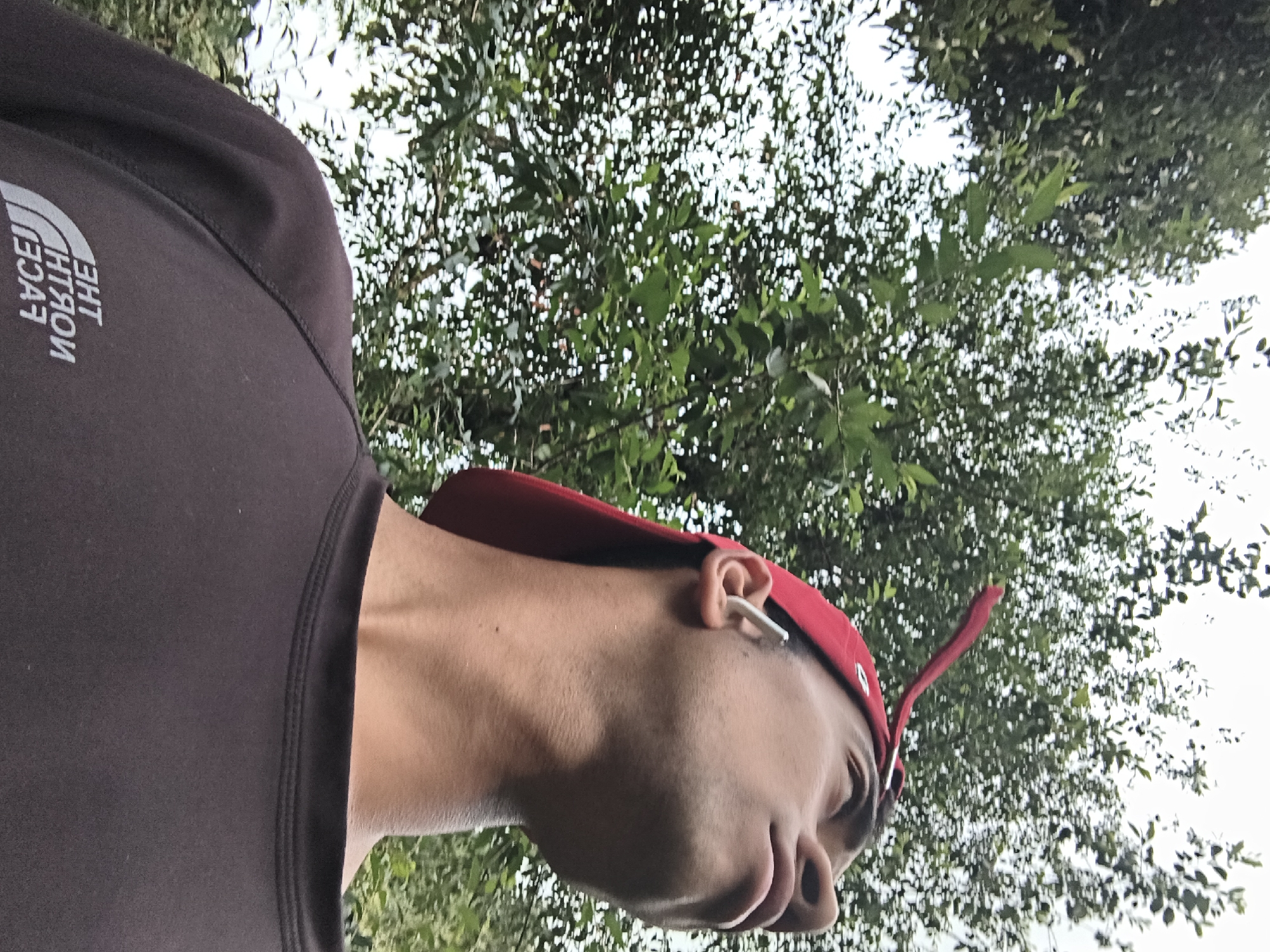 A man wearing a brown sweatshirt with "THE NORTH FACE" written on the sleeve and a red baseball cap is positioned in front of a tree with dense green foliage. An earbud is visible in his ear. The background appears to be a sky obscured by the tree canopy. The image orientation is rotated. No other details about Haini, India are present.