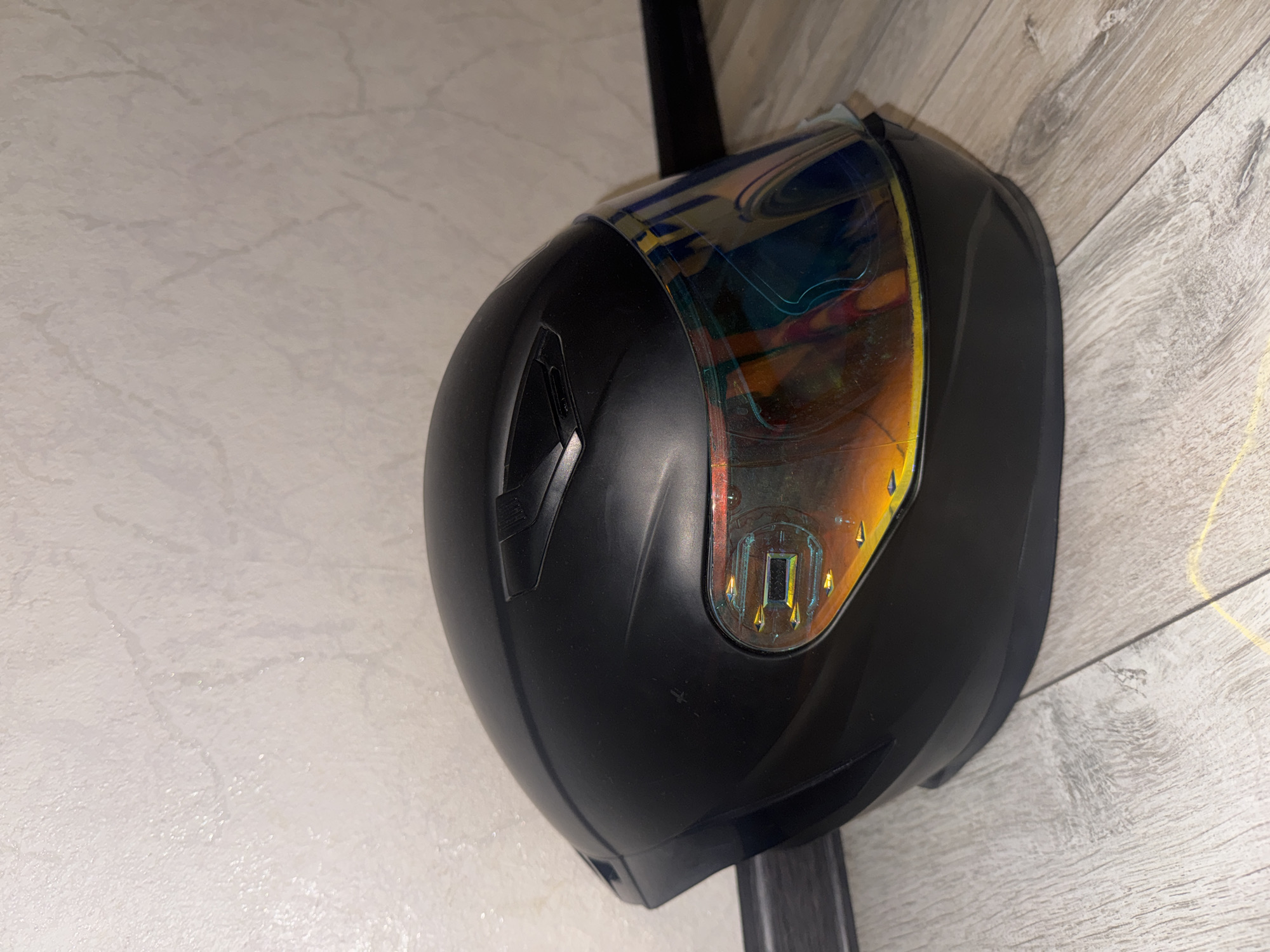 A black motorcycle helmet with a multi-colored reflective visor rests on a light-colored wooden floor in front of a textured, off-white wall. The helmet is positioned at an angle, with the visor facing the viewer and reflecting light in hues of orange, yellow, blue, and green. The wall appears to have a subtle, irregular pattern, possibly from wallpaper or a painted finish. A dark, narrow strip, likely a baseboard or furniture edge, runs horizontally along the bottom of the wall. The scene is indoors, with the lighting suggesting a domestic or retail environment. There are no people or animals present in the image.