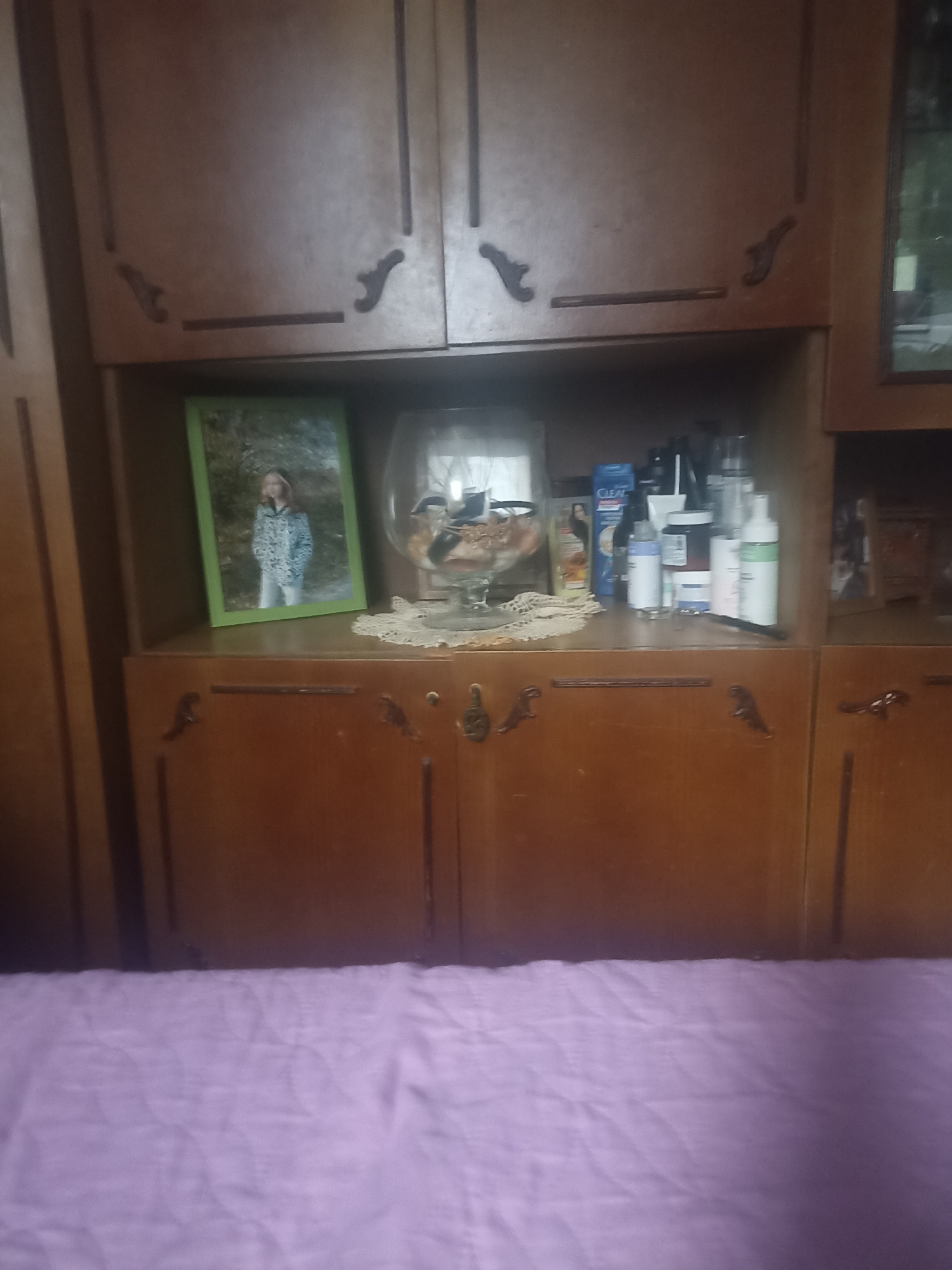 An indoor scene depicts a wooden cabinet with two visible doors on the lower half and two visible doors on the upper half. The upper cabinets have decorative carvings. The lower cabinet doors have ornate handles. The open shelf above the lower cabinets displays a framed photograph of a young girl, a glass bowl with decorative items, and several bottles of toiletries. To the right of the shelf, a framed picture is visible within a cabinet. The scene is viewed from a lower vantage point, with a creased lavender sheet or blanket occupying the bottom portion of the image. The location is identified as Perelešinskij, Russia.