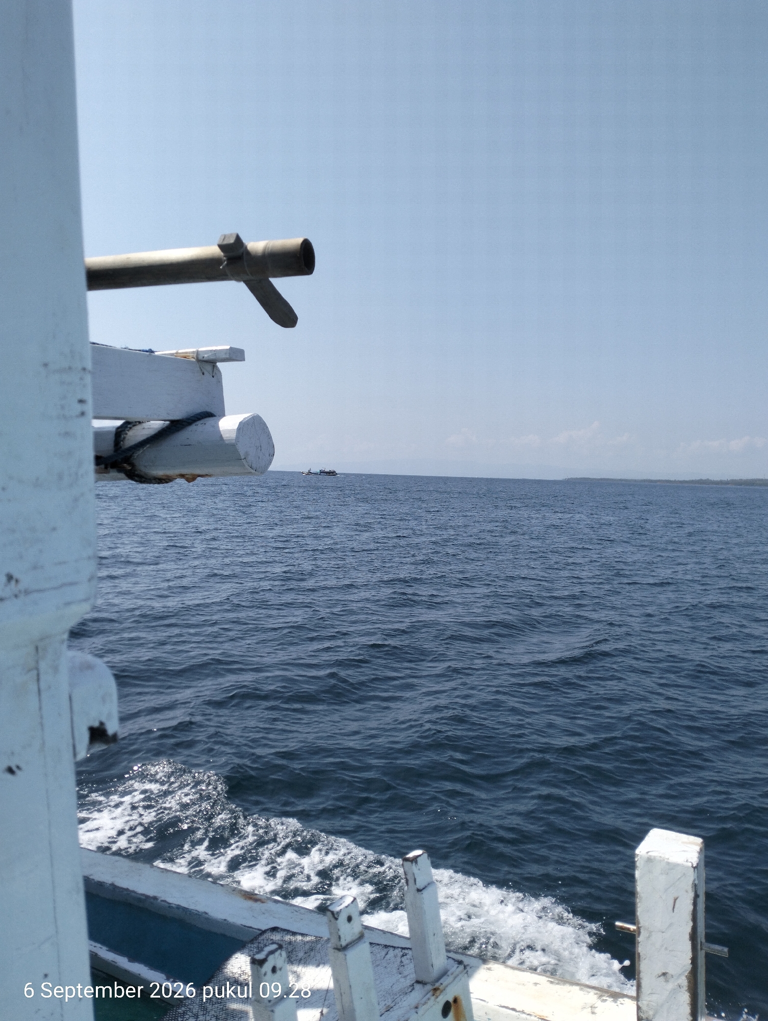 The image shows a view from a boat at sea, with the dark blue water stretching to the horizon under a clear sky. In the foreground, the white painted side of the boat is visible, with a metal pipe and wooden fixtures. The water is disturbed, creating white foam and ripples on the surface, indicating movement. In the distance, two small boats can be seen on the water, and a faint line of land is visible on the horizon. The timestamp "6 September 2026 pukul 09.28" is overlaid in the bottom left corner. The scene is likely a nautical setting near the city of Kendari, Indonesia, possibly in the waters of the Banda Sea or near the coast of Sulawesi. There are no people visible in the image.