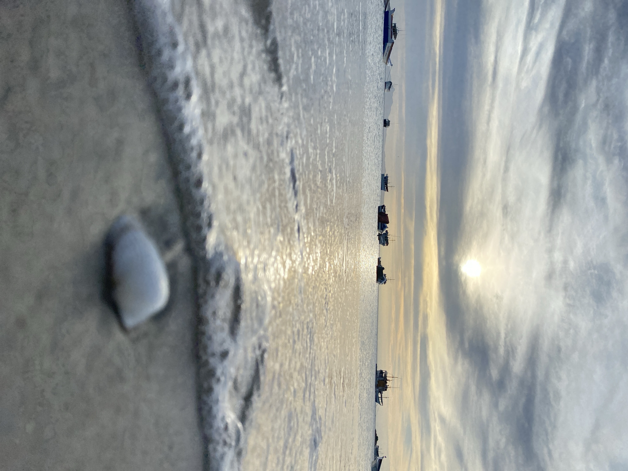 The image captures a serene coastal scene in Juan Griego, Venezuela. In the foreground, a small, light-colored seashell rests on the wet sand. The tide is coming in, with gentle waves lapping at the shore. The water reflects the sunlight, creating a shimmering effect. Further out in the water, several boats are anchored, their silhouettes visible against the horizon. The sky is filled with clouds, with the sun peeking through, casting a warm glow over the scene.