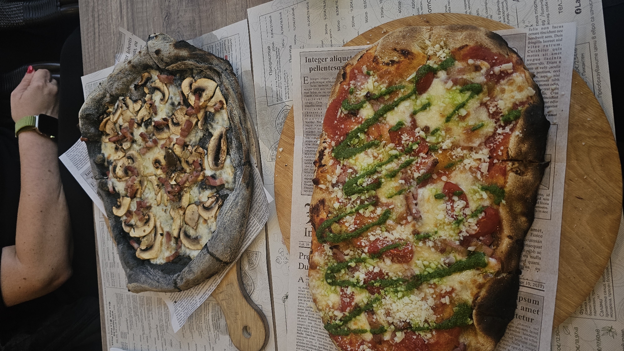 Here's a detailed description of the image:
**Scene Overview:** The image features two pizzas, each on a separate wooden serving board, presented on a table covered with newspaper-style placemats. A person's arm and hand, wearing a green Apple Watch, are visible on the left side of the frame.
**Subjects:**
* **Pizzas:** The pizza on the left has a dark, possibly charcoal-infused crust, topped with cheese, mushrooms, and bacon. The pizza on the right has a lighter crust with a tomato sauce base, cheese, tomatoes, bacon, and green pesto drizzle.
* **Person:** A person is visible to the left with a green smartwatch and reddish nail polish.
**Objects:**
* **Wooden Boards:** Each pizza rests on a separate wooden serving board.
* **Placemats:** The table is covered in placemats with a newspaper print design.
* **Watch:** A green Apple Watch is visible on the person's wrist.
**Actions/Interactions:**
* The scene suggests a meal being served and photographed at a restaurant.
**Environment:**
* The scene is likely set in a restaurant. The presence of the placemats suggests it's a casual dining establishment.
* The text on the placemats mentions "www.lapala.bg" and "La Pala Pizza". Also, there is an address listed as "+359 877 66 55 58" indicating the establishment is likely in Bulgaria.
**Location and City:**
* Based on the URL and telephone number, the location can be tentatively identified as being in Bulgaria. It is likely from a restaurant named "La Pala Pizza" which is located in the city of Varna, Bulgaria.