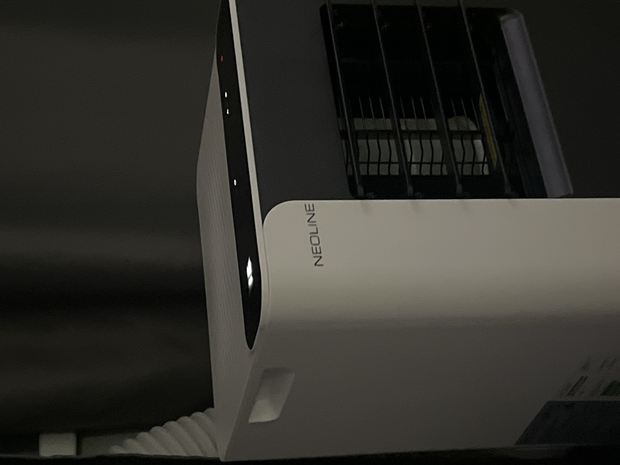 The image shows a close-up of a white electronic device with the brand name "NEOLINE" visible in dark text. The device is dark gray on top, with a black grille-like section. There are several small indicator lights on the side of the white portion, some of which are lit. The device appears to be a dashcam or similar automotive electronic. The background is dark and out of focus, suggesting the photo was taken indoors, possibly in a car or a dimly lit room. The lighting is low, and the overall mood is neutral. No specific time of day or weather conditions can be inferred. The location context provided is Krasnoyarsk, Russia.