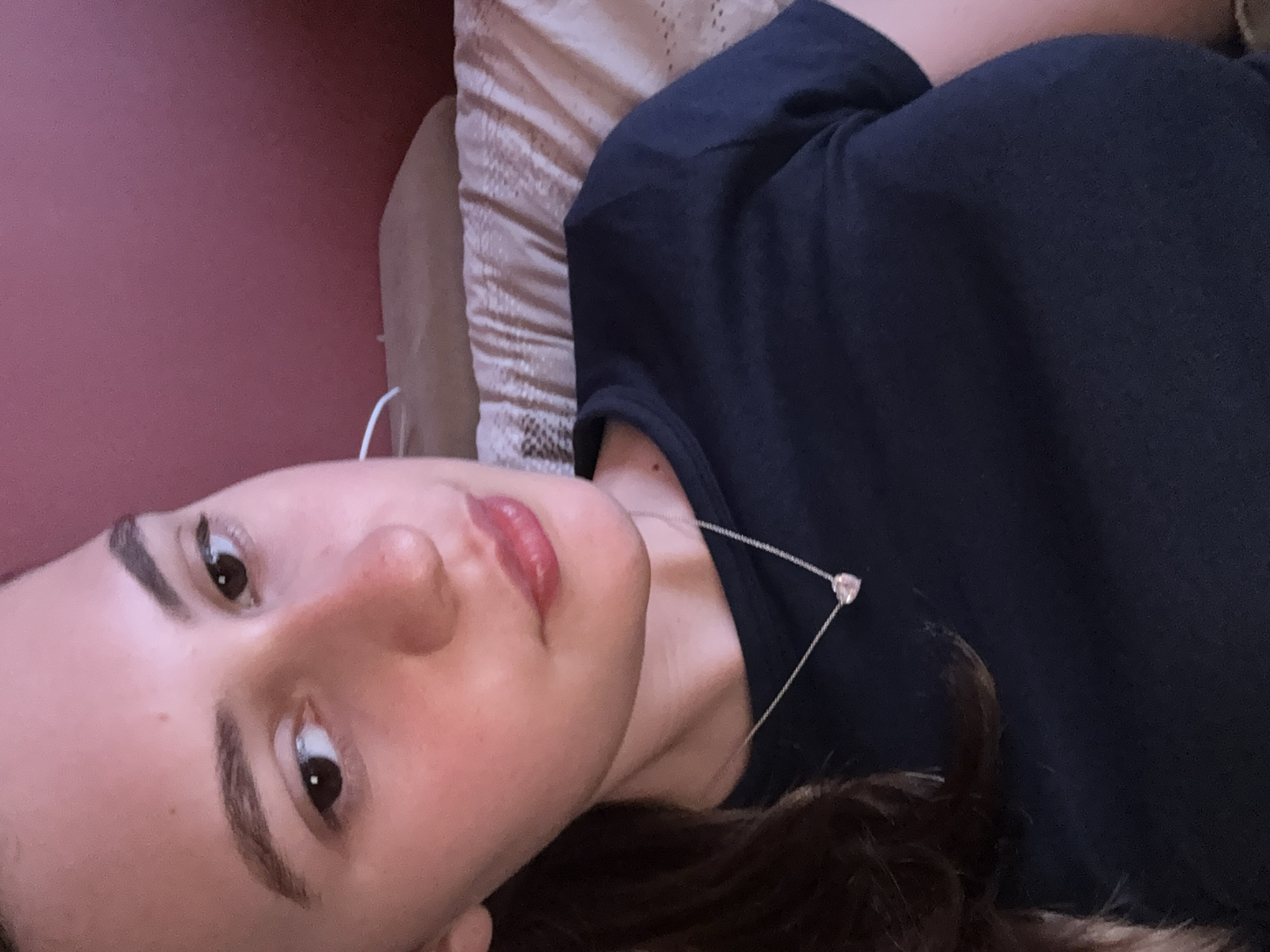 A young woman with dark brown hair is lying down. Her head is tilted to the left. She is wearing a dark blue T-shirt and a silver necklace with a heart-shaped pendant. The background includes a pink wall and a beige patterned pillow. The location is undetermined, with no information provided to suggest Panagyurishte, Bulgaria.