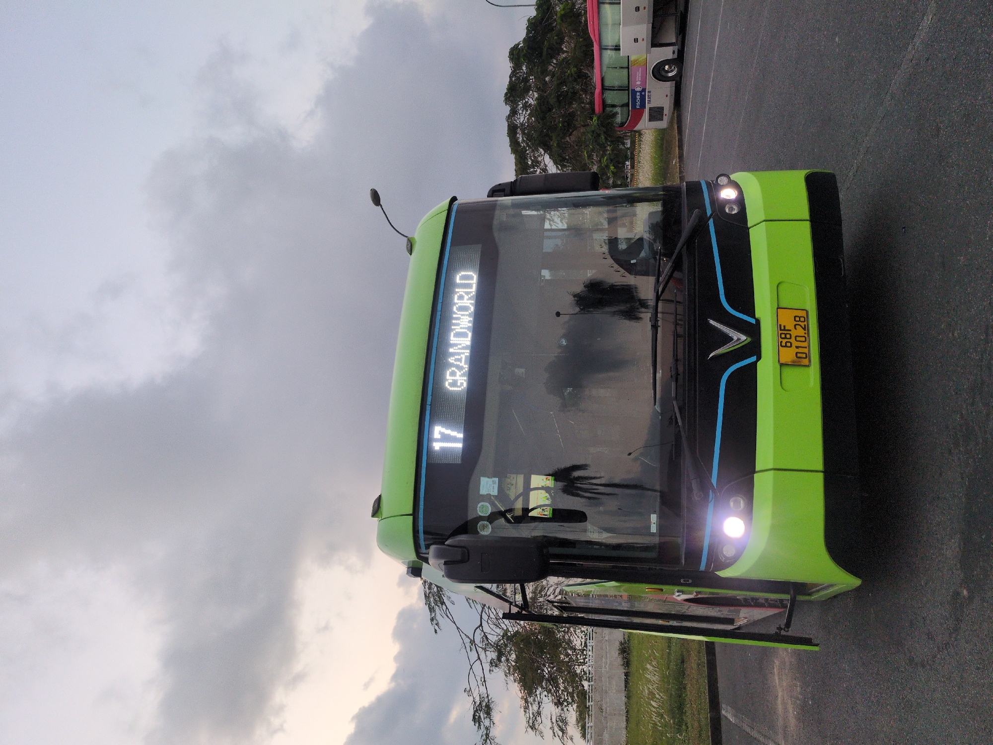 The image shows a green bus with the number "17" and the destination "GRANDWORLD" displayed on its front. The license plate reads "68F 010.28". The bus is on a road, and another bus is partially visible in the background. Trees are visible in the background, and the sky is overcast. Based on the license plate code "68F," the bus is located in the city of Phú Quốc, Vietnam. The presence of the "GRANDWORLD" destination suggests the bus is traveling to or from the Grand World Phu Quoc complex. The bus's door is open, indicating it may be stopped or about to start its route. No people are visible in the immediate vicinity of the bus.
