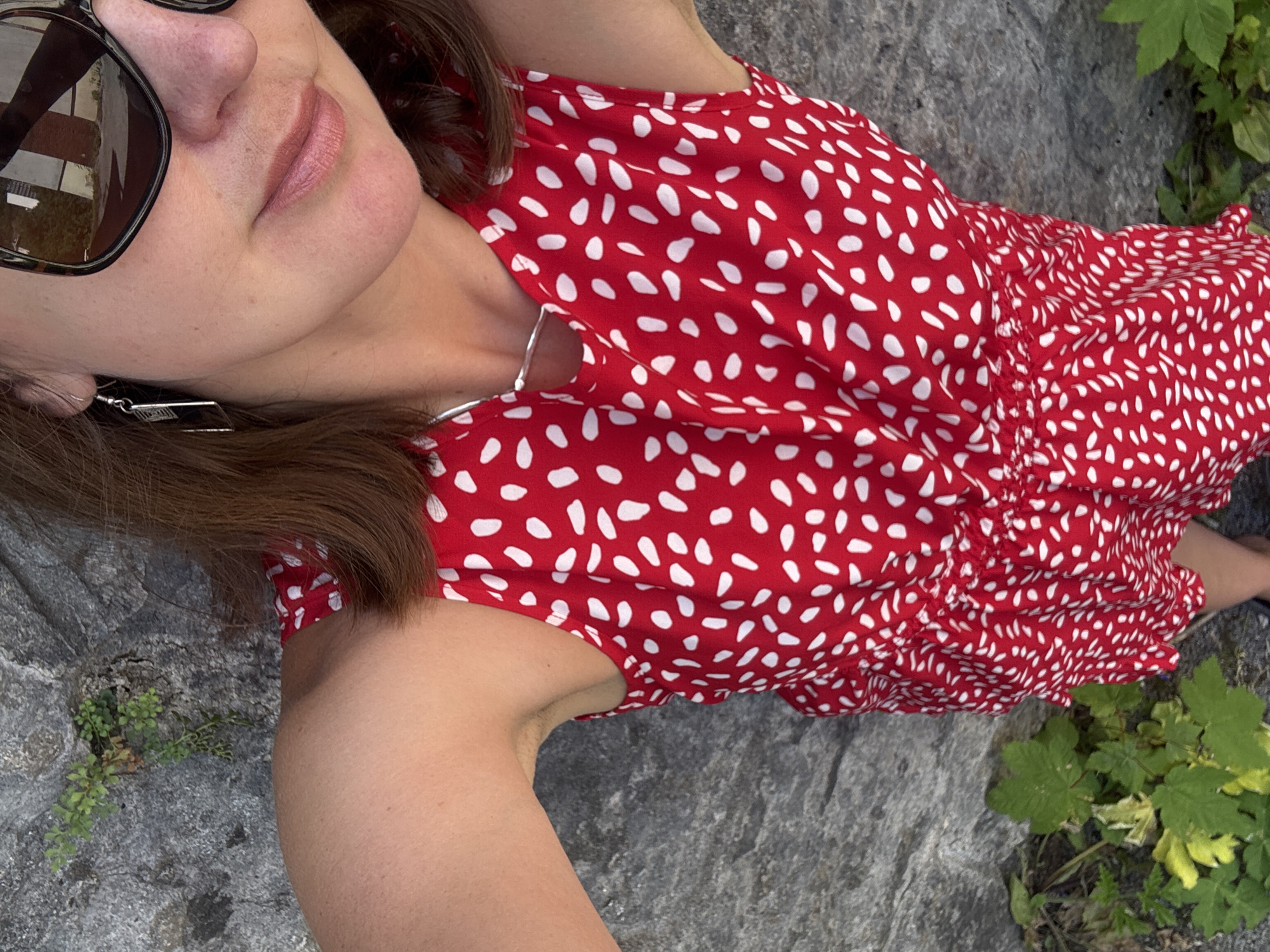 A woman is captured from a high-angle, selfie-style perspective outdoors. She is wearing a red and white polka dot dress and large, dark sunglasses. Her right arm is raised, holding the camera. The background consists of a rough, grey stone wall and some green foliage. The overall impression is one of a casual outdoor setting, possibly in a park or natural area within the city of Saint-Claude, France.
