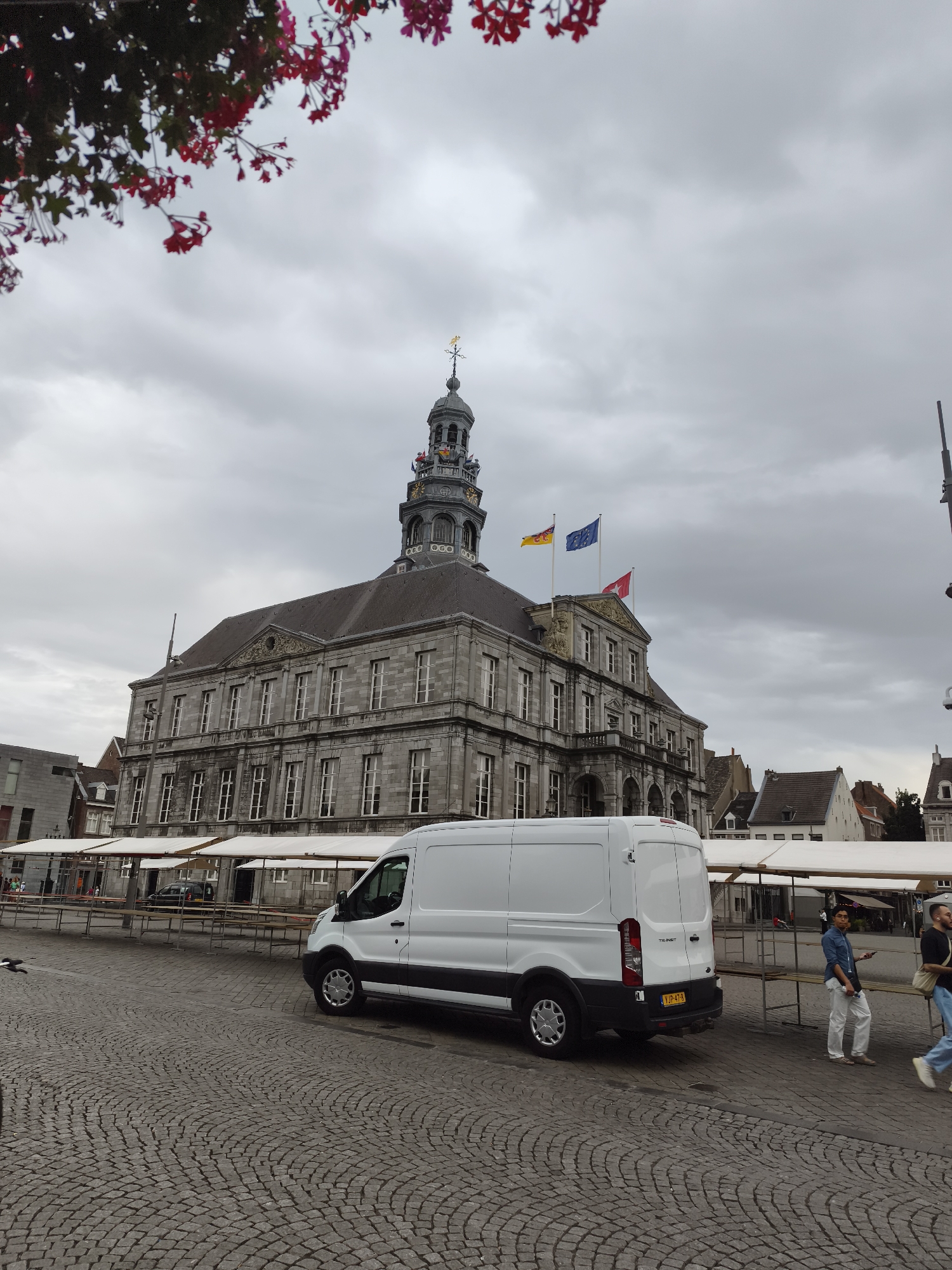 The image depicts the Markt square in Maastricht, Netherlands, dominated by the stately facade of the Maastricht City Hall. A white Ford Transit van is parked on the cobblestone square, its rear facing the camera. Several market stalls with white canopies are set up in front of the City Hall. Two young men, one in a blue shirt and white pants, the other in a dark shirt and jeans, are walking across the square. The sky is overcast with gray clouds. Overhead, pink flowers are visible, possibly from a tree or hanging baskets. The City Hall features a prominent clock tower with flags flying from its upper levels. The architectural style of the City Hall is classical, with stone construction and numerous windows.