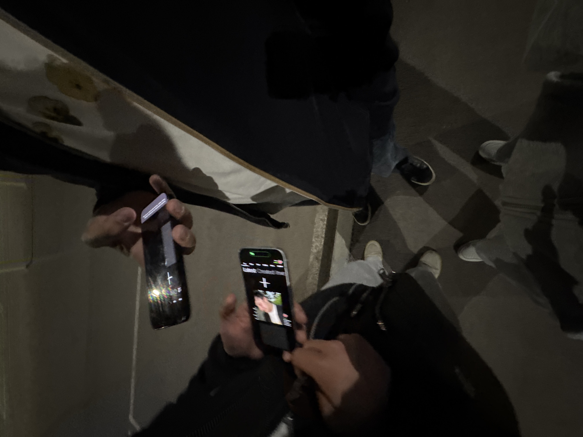 The image captures a nighttime outdoor gathering in Yerevan, Armenia, with a focus on people interacting with their smartphones. In the foreground, two individuals hold their phones, both displaying illuminated screens. The central phone prominently features a screen showing what appears to be a video or photo of a person, with a '+' icon and barely legible text that seems to indicate "current" content or a caption. A green battery indicator is visible at the top of this phone's display. The second phone, held to the left, also shows screen reflection, suggesting it's active.
The scene is dimly lit, emphasizing the artificial light from the phone screens. Elements of people's attire are visible, including dark sleeves, jeans, and light-colored sneakers on pavement, indicative of a casual setting. In the upper left portion of the image, an off-white, board-like object, possibly the underside of a skateboard, with circular markings, is partially visible. The overall atmosphere suggests a casual social interaction among friends or acquaintances, likely sharing content or engaging with their devices in the evening.