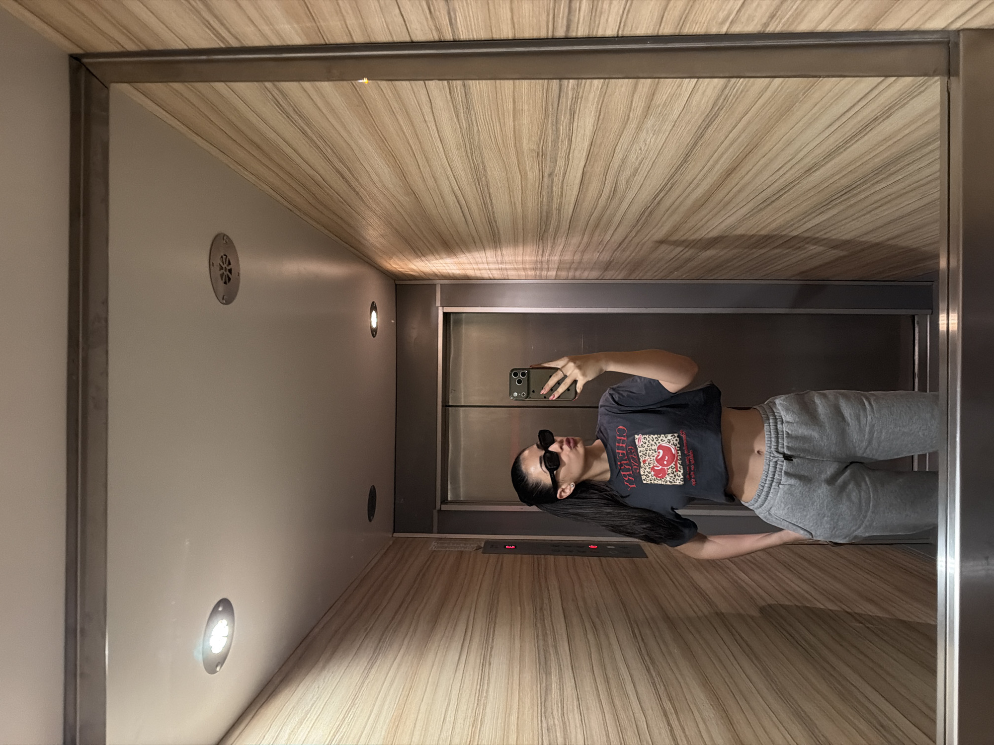 The image depicts a person standing in an elevator, taking a selfie with a smartphone. The elevator's interior features wood-paneled walls and ceiling, along with metal accents on the doors and frame. The person is wearing sunglasses, a dark cropped t-shirt with a graphic, and gray sweatpants. There are lights and ventilation elements visible on the elevator walls. It is not possible to determine the location in Sofia, Bulgaria from the given image alone.