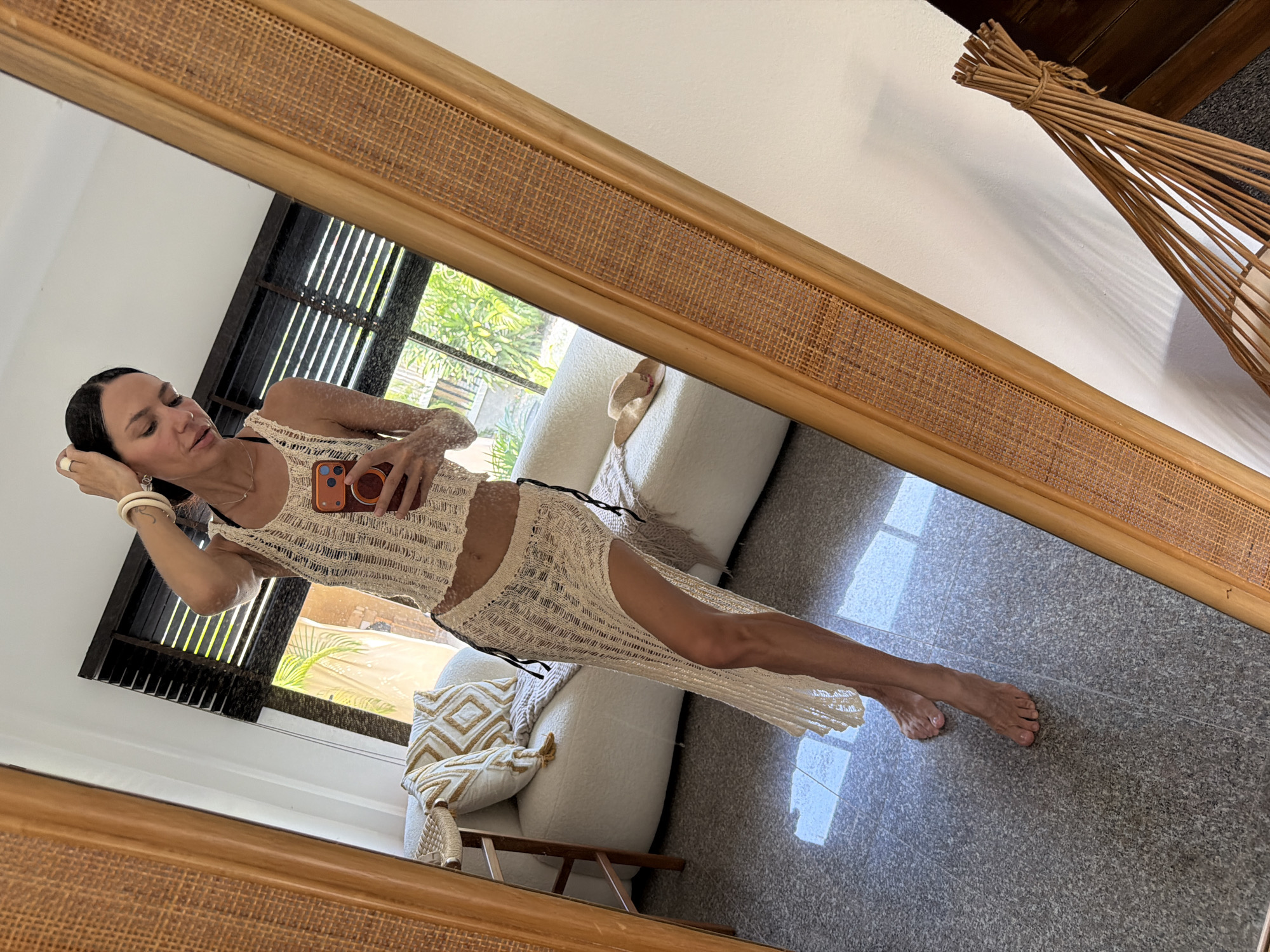 A woman stands in front of a full-length mirror in what appears to be a well-lit room. She is wearing a cream-colored crochet two-piece outfit, consisting of a sleeveless crop top and a long, flowing skirt with a high slit. The outfit is see-through, revealing a darker bikini underneath. She has her hair pulled back and is holding her phone in her right hand, taking a selfie. Her left hand is raised to her head, and she is wearing bracelets and a ring.
The background reflected in the mirror shows a modern living space with a plush white sofa adorned with decorative pillows. A straw hat rests on the sofa. Sunlight streams through blinds, casting shadows on the floor. The room has a tiled floor and a large mirror with a wooden and woven bamboo frame. To the right of the mirror, a decorative object made of bundled bamboo sticks is visible.
The overall impression is of a casual, resort-like setting, possibly a hotel room or private residence, in the city of Baan Bo Phut, Thailand, given the ambient light and greenery visible through the window.
