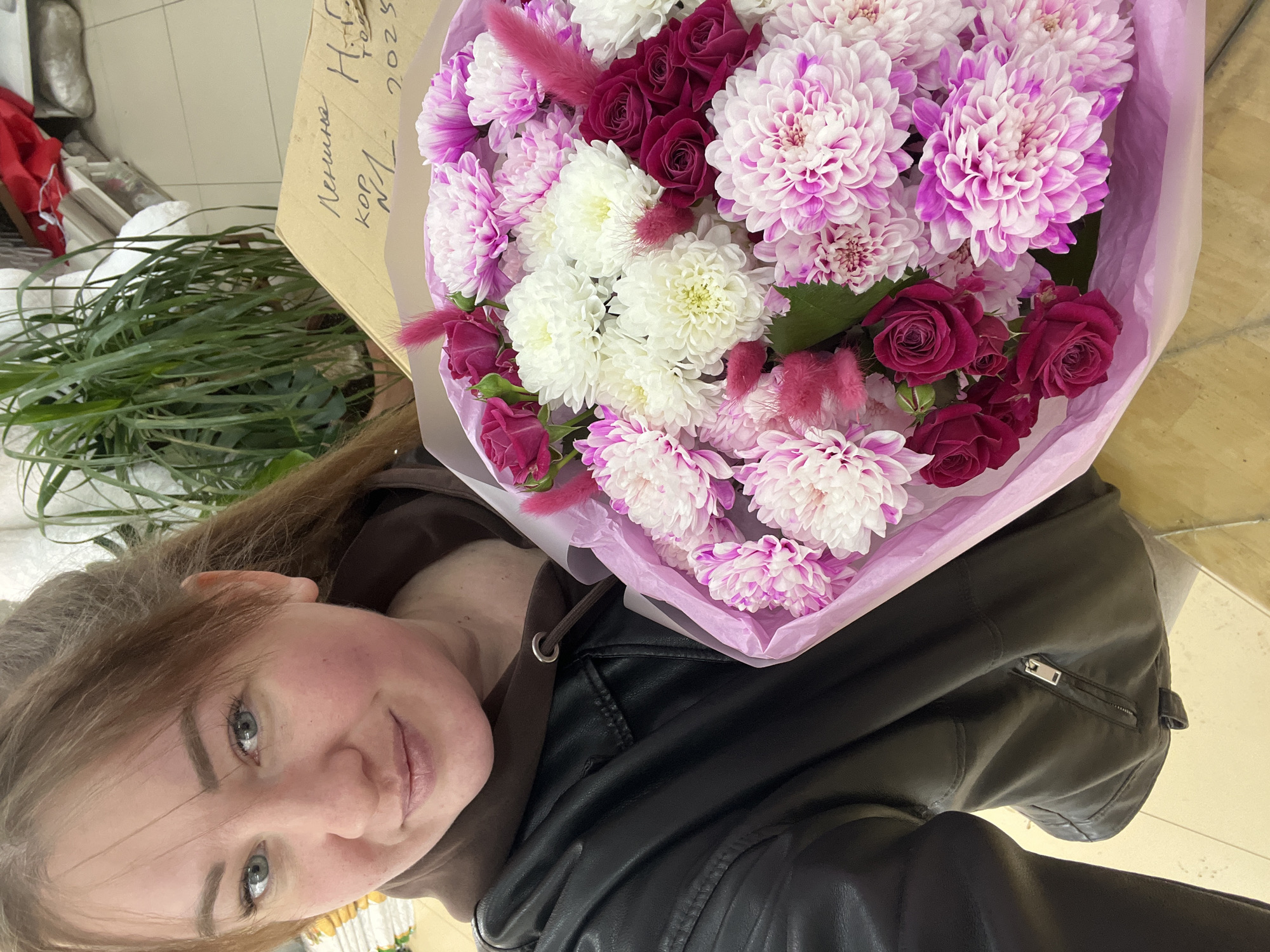 A woman with light brown hair and light blue-green eyes is depicted in a chest-up selfie, holding a large flower bouquet. She wears a brown hooded sweatshirt under a black leather jacket and smiles subtly towards the camera. The bouquet, wrapped in light purple paper, contains white chrysanthemums, two-tone pink and white dahlias/chrysanthemums, small deep pink spray roses, and fluffy pink decorative elements. The scene is indoors, featuring a light-colored tiled floor. In the background, a brown cardboard box displays handwritten Cyrillic text, including "Ленина" (Lenina), "кор." (korpus/building), and "2025", indicating an address on Lenina Street in Chaikovsky, Russia. A large potted plant with long green leaves is visible behind the box, alongside other indistinct items on shelves.
