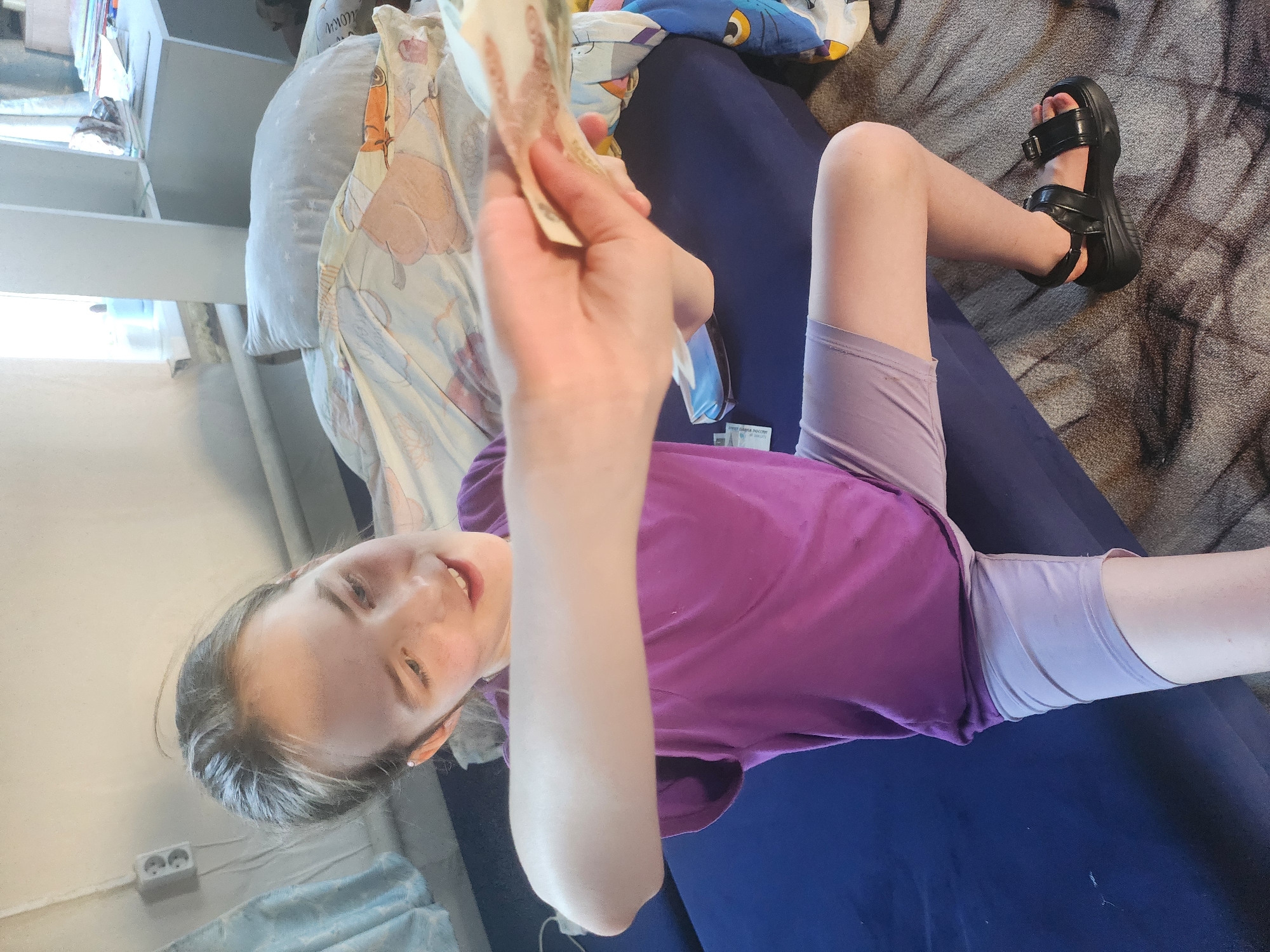 A child is sitting on a blue surface, possibly a sofa or bed, in what appears to be an indoor setting. The location is likely in Krasnogorskoye, Russia. She is wearing a purple t-shirt and light purple shorts. She is holding Russian currency in her hand, presumably rubles. On her feet are a pair of black platform sandals. In the background, there is a patterned pillow, and a shelving unit is visible on the left side of the frame. A gray, patterned rug is present in the upper right corner of the image. The child is looking towards the camera with a slight smile.