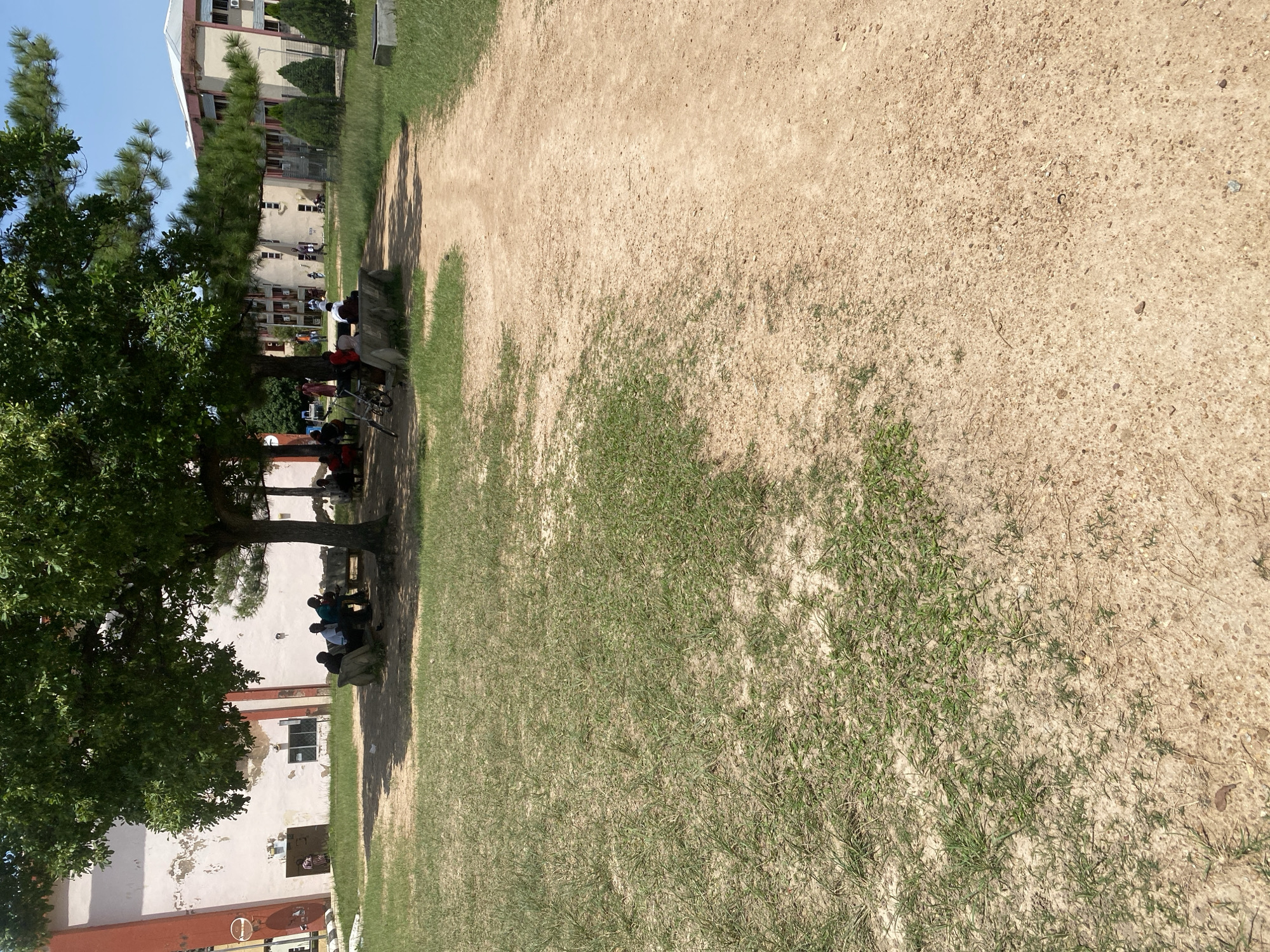 The image depicts a group of people gathered under the shade of a large tree in a park-like setting. A dirt path runs through the scene, leading towards buildings in the background. The buildings appear to be part of an institutional or residential complex, with multiple stories and windows visible. The overall environment is outdoors, with grass and sparse vegetation. Several individuals are seated on benches or on the ground beneath the tree, engaged in conversation or resting. The scene suggests a relaxed, communal atmosphere, possibly during a break or leisure time. The architecture of the buildings and the presence of mature trees hint at a location in Jos, Nigeria, a city known for its plateau topography and cooler climate.