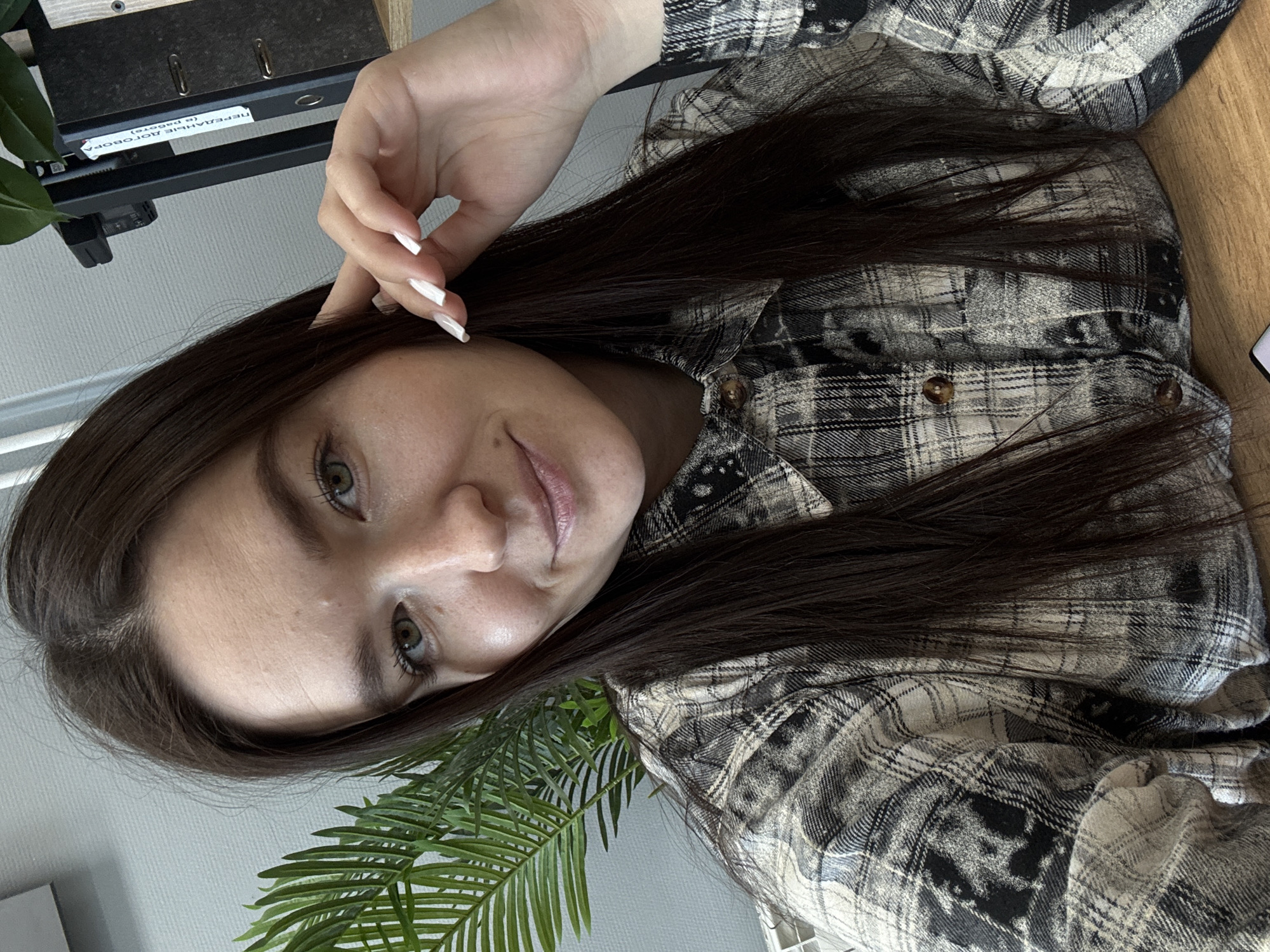 A selfie of a young woman is presented. Her dark brown, straight hair cascades across her shoulder and around her face, framing her light skin and prominent cheekbones. She has fair skin and light-colored eyes. Her hand, with long, white nails, is positioned near her temple. She wears a gray and black flannel shirt. A green, artificial palm plant is in the lower left corner. In the background, a wall and a document holder with paperwork are visible. The location is possibly indoors, in a personal or office space, but the image does not reveal specific details indicating it is in Chaikovsky, Russia.