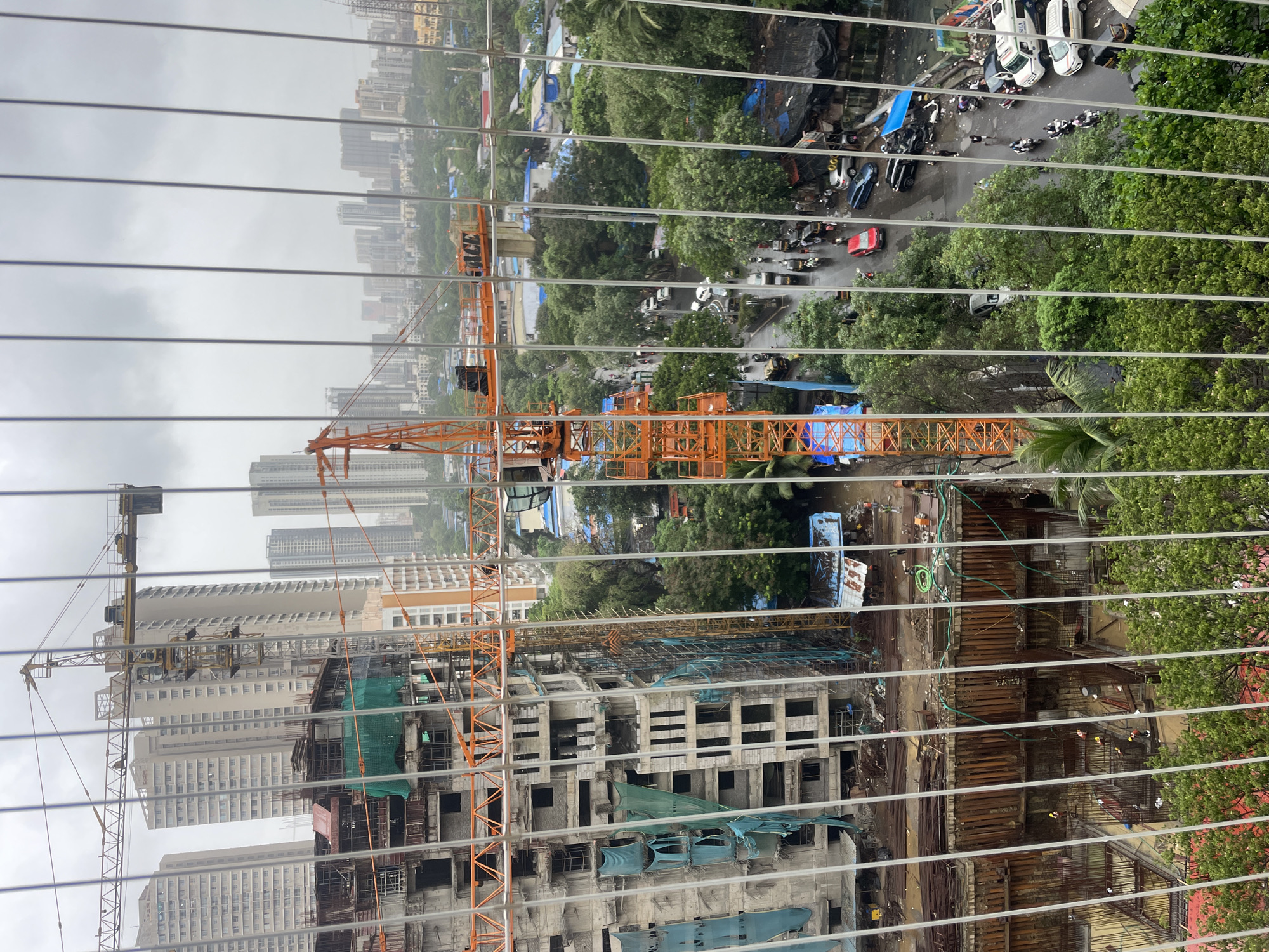 This is an aerial, eye-level view of an urban construction site in Mumbai, India, on a cloudy day. A large, orange tower crane dominates the center of the frame, positioned next to a multi-story building under construction. The building has exposed rebar and concrete, with blue tarps draped over some sections. In the foreground, a wooden scaffold structure is visible. Beyond the construction site, a city skyline with numerous high-rise buildings can be seen. A street runs through the middle ground, with cars and motorcycles driving on it. Lush green trees are interspersed throughout the scene. The image is partially obscured by thin, vertical white wires, likely from a balcony railing. The overall atmosphere suggests a busy construction project in a densely populated urban environment.