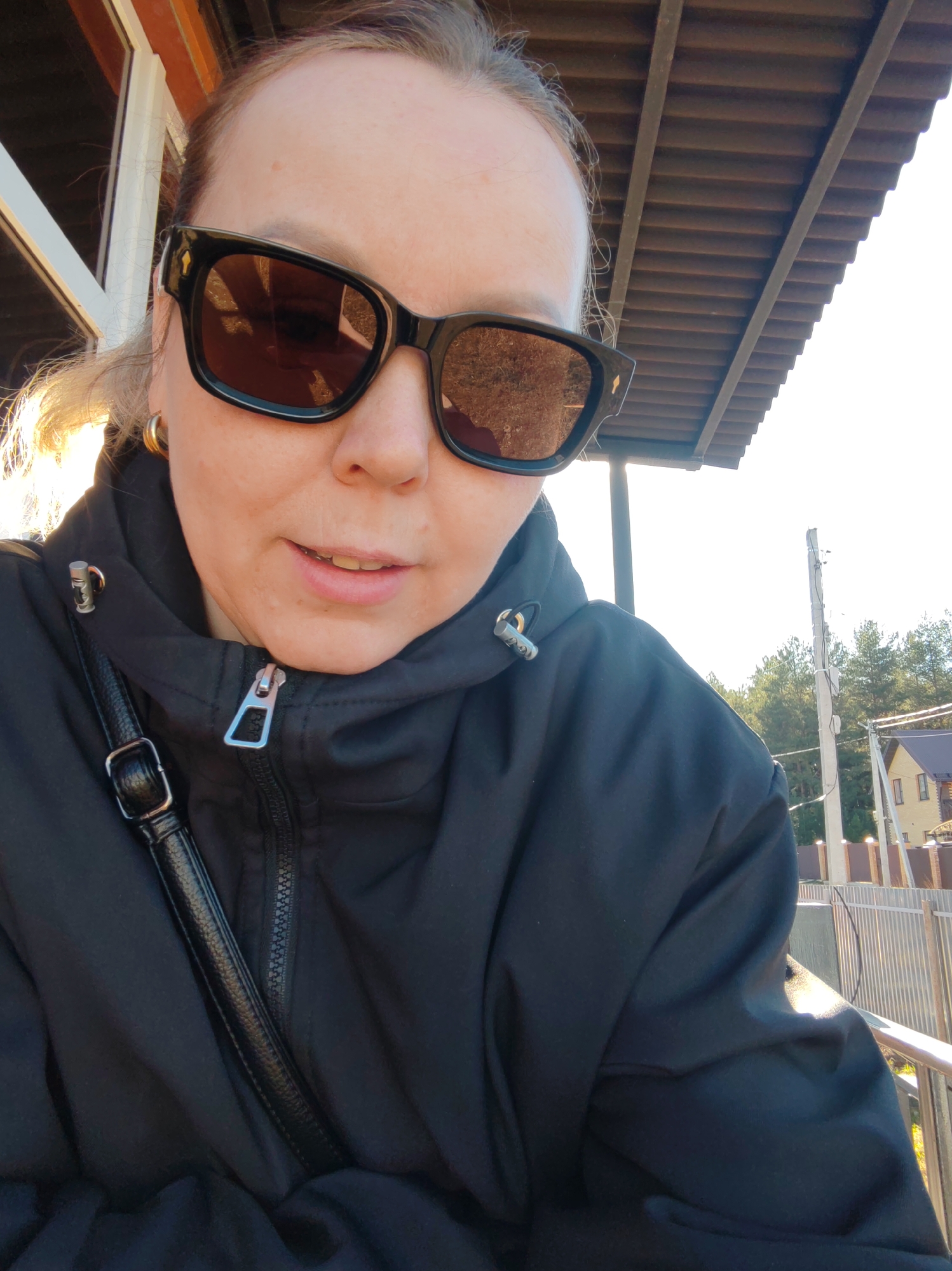 A woman in a black jacket and sunglasses is posing for a selfie. She is standing under a roof structure. A black purse strap is visible across her chest. The background includes a power pole, trees, and a house. The setting appears to be Chaikovsky, Russia.