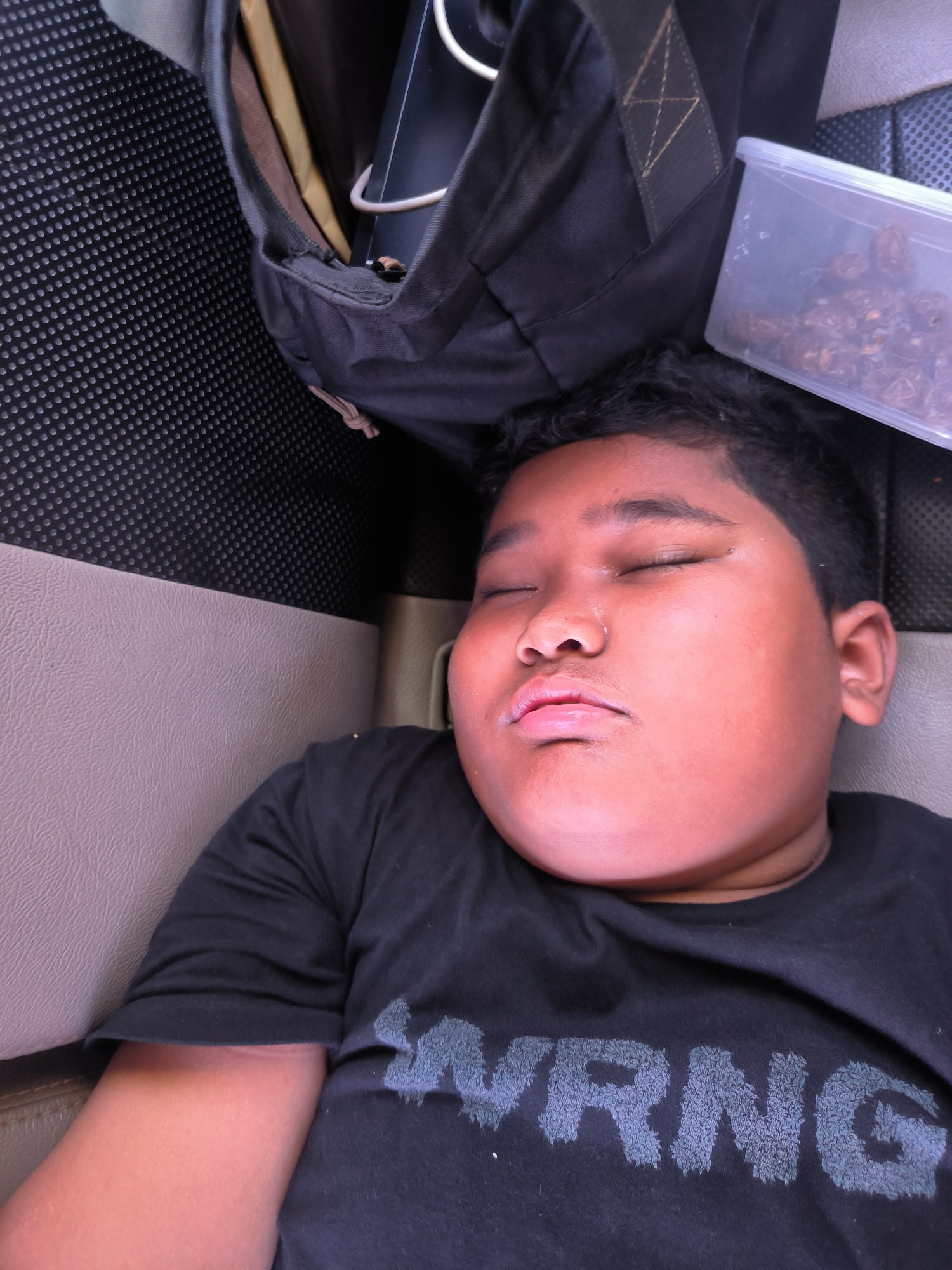 The image displays a close-up, slightly overhead view of a young child sleeping in what appears to be the interior of a vehicle, possibly in Banda Aceh, Indonesia. The child, with a tan complexion and dark, short curly hair, is shown from the chest up, resting with eyes closed and mouth slightly open, revealing a bit of saliva on the lower lip. They are wearing a black t-shirt with the partial text "WRNG" visible on the chest in a dark, textured font.
The setting suggests an automobile or bus interior, with a beige-colored seat or panel forming the background beneath the child's head and a dark, textured material (possibly a floor mat or seat back) above. A dark-colored backpack is partially visible in the upper left, containing some items including a white cable. To the right of the backpack, a clear plastic container filled with dark, dried fruit, resembling raisins or similar berries, is present. The lighting is diffused, consistent with an indoor or vehicle environment, making the specific time of day or external weather conditions unclear. The child's posture and closed eyes indicate they are asleep, likely resting during a journey.