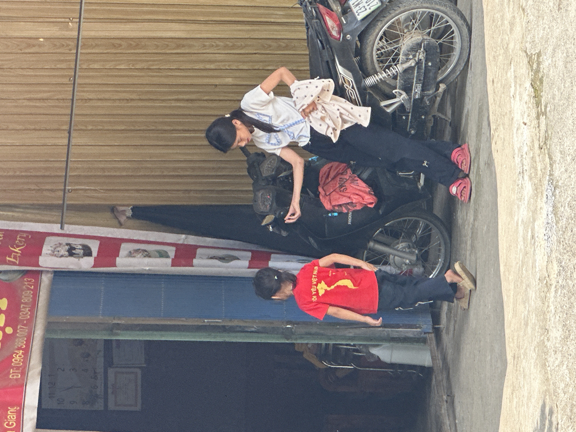 This image captures a candid moment on a sunny day in Du Già Commune, Vietnam, outside a shop. Two individuals are featured: an adult female (or older girl) and a child.
The adult, with dark hair tied back, is wearing a white short-sleeved top with blue embroidered details and dark pants, paired with red sandals. She is leaning over a parked black motorcycle, possibly tending to something on its seat, where a red fabric item is placed. She holds a light-colored polka-dotted cloth in her hands. Next to her, another black motorcycle is visible.
The child, wearing a red t-shirt emblazoned with the Vietnamese phrase "TÔI YÊU VIỆT NAM" (I LOVE VIETNAM) and a yellow map of Vietnam, stands near the shop's entrance. The child also wears dark pants and light-colored sandals.
The background features the exterior of a building with a light brown, wooden-slat upper facade and a closed blue roll-down door below it. To the left, a red and white banner advertises a "Bakery" and includes phone numbers: "ĐT: 0964 368 007 - 0347 809 213". Through a glass door or window below, a wall clock inside the shop is visible, showing the time as "11:12". The motorcycles bear partial license plates, with "23-M1" and "21" discernible on one, and a "HONDA" logo is also visible. The ground is a light-colored concrete pavement. The bright daylight suggests it's late morning or midday.