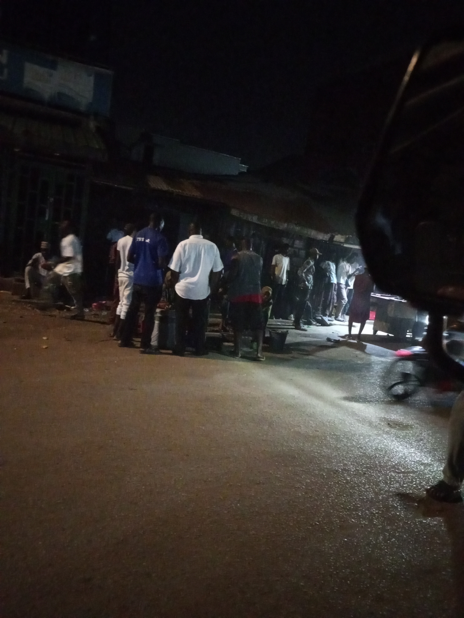 A nighttime scene in Maiburiji, Nigeria shows a group of approximately 15 people gathered along a street lined with shops or stalls. Most individuals are standing, wearing casual clothing such as t-shirts and trousers. Some are interacting with others in the group. Some are facing the street. Street lighting illuminates the wet pavement, suggesting recent rain. The scene is framed by the side of a vehicle or object, appearing large and dark in the foreground.