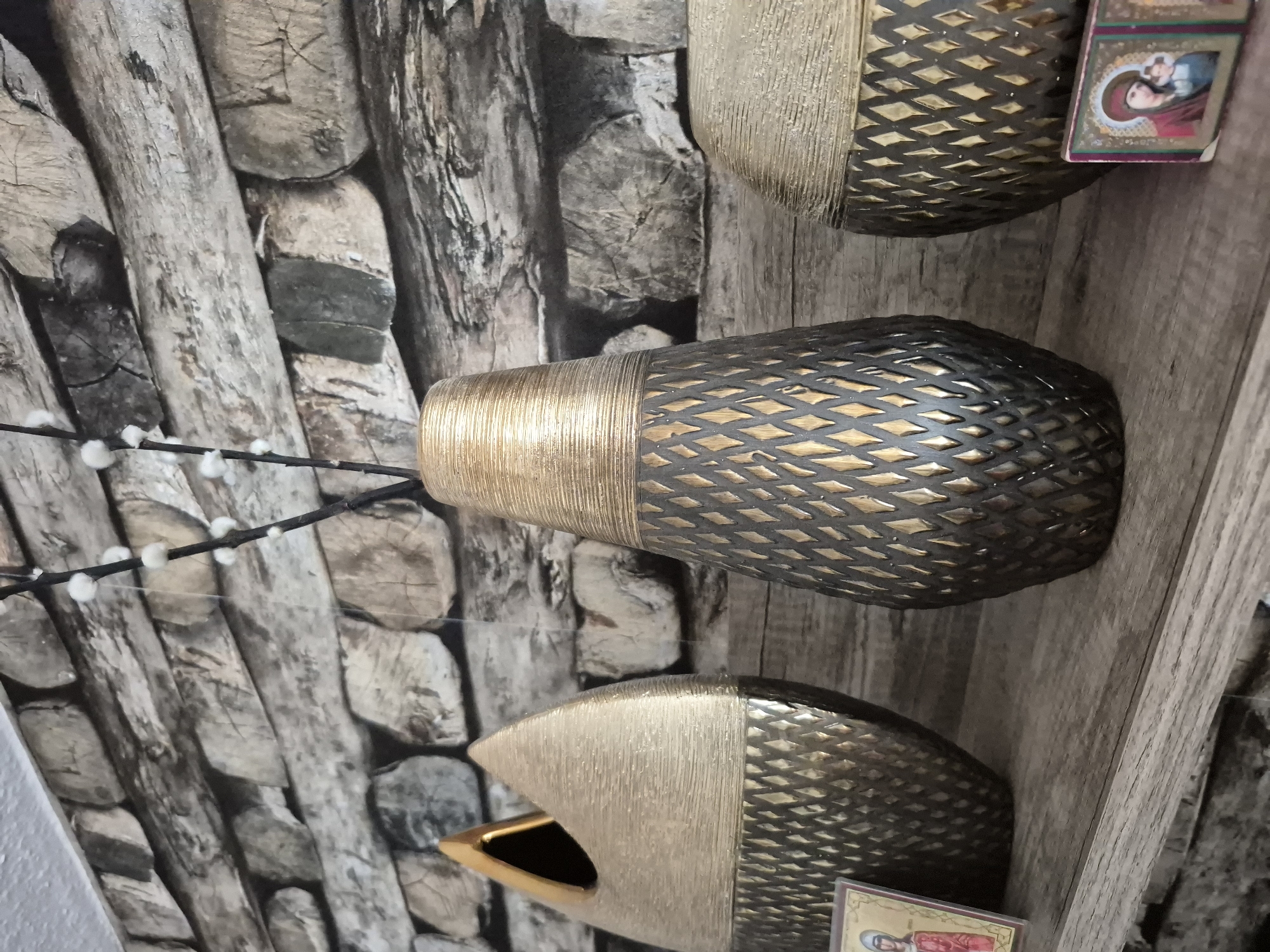 A close-up, eye-level shot showcases a shelf decorated with three ornate vases and two religious icons. The backdrop features a textured wallpaper resembling stacked logs. The vases, varying in size and shape, are predominantly gold and dark gray, with intricate diamond-patterned designs. One vase is a tall, slender urn with a ribbed gold top and a cutout diamond pattern. Another is a wider, squat vase with a similar ribbed gold top and diamond pattern. The third vase has a unique, asymmetrical teardrop shape with a gold interior and a textured gold and gray exterior. Placed to the left of the leftmost vase is a small, rectangular religious icon depicting a woman with a child. To the right of the rightmost vase, another, similar religious icon is visible. The lighting is soft, casting subtle shadows. The scene is static, with no people or animals present.