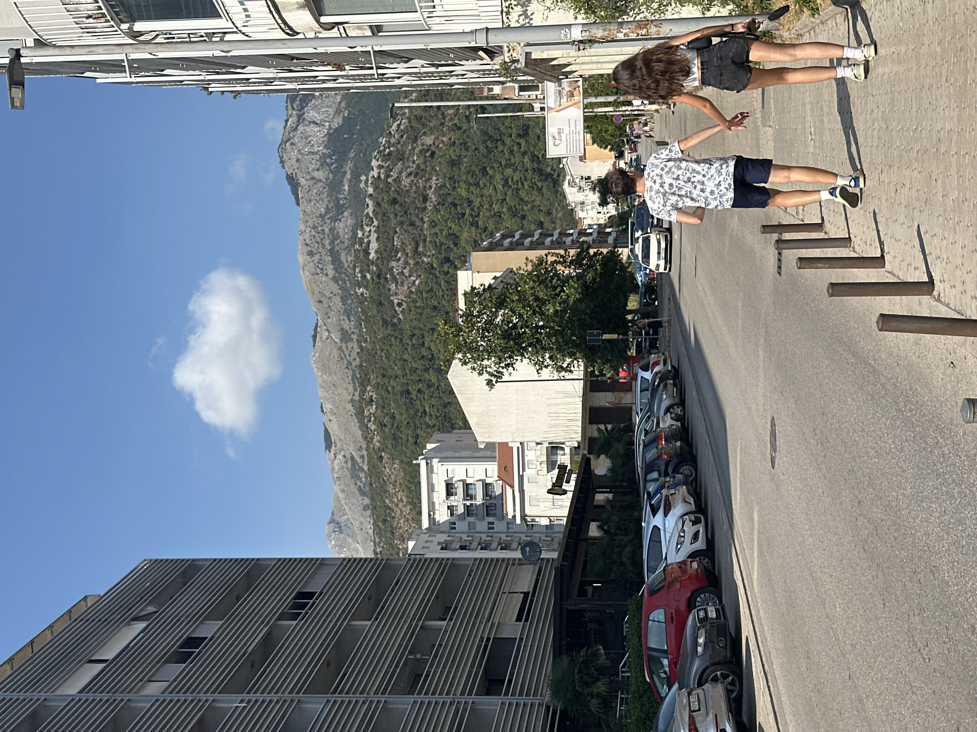 The image shows a street scene in Budva, Montenegro, under a clear blue sky with a single fluffy cloud. On the left, a modern, multi-story building with horizontal slats dominates the foreground. Across the street, parked cars are lined up along the curb. In the mid-ground, various buildings of different architectural styles are visible, including apartment complexes and what appears to be commercial establishments. Further in the background, a lush, green mountainside rises, dotted with trees, and its rocky peaks are visible against the sky.
In the foreground, a young couple is walking hand-in-hand on the sidewalk, facing away from the camera and towards the street. The male is wearing a patterned t-shirt and dark shorts, and the female is wearing a white top and black shorts. They are walking on a paved sidewalk with metal bollards along its edge. The street itself is asphalt, with a yellow taxi visible in the distance, along with other vehicles. The overall atmosphere suggests a warm, sunny day in an urban environment with a scenic natural backdrop.