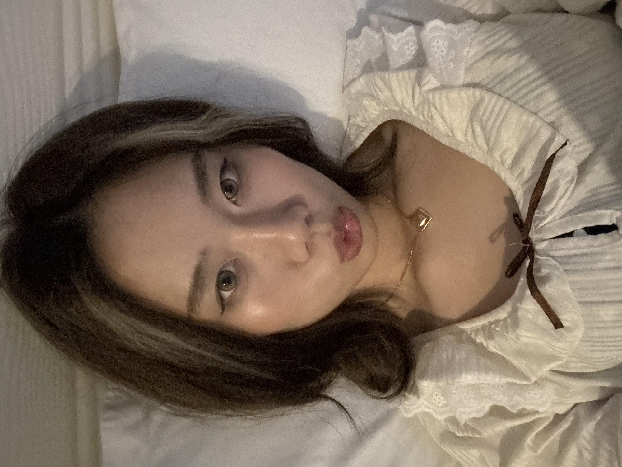 The image shows a woman lying on a white bed in what appears to be a bedroom. She has brown hair with lighter highlights and is wearing a white, ruffled shirt with a brown ribbon. She is wearing a gold necklace with a square pendant. The location cannot be determined from the image, nor the actions, or interactions.