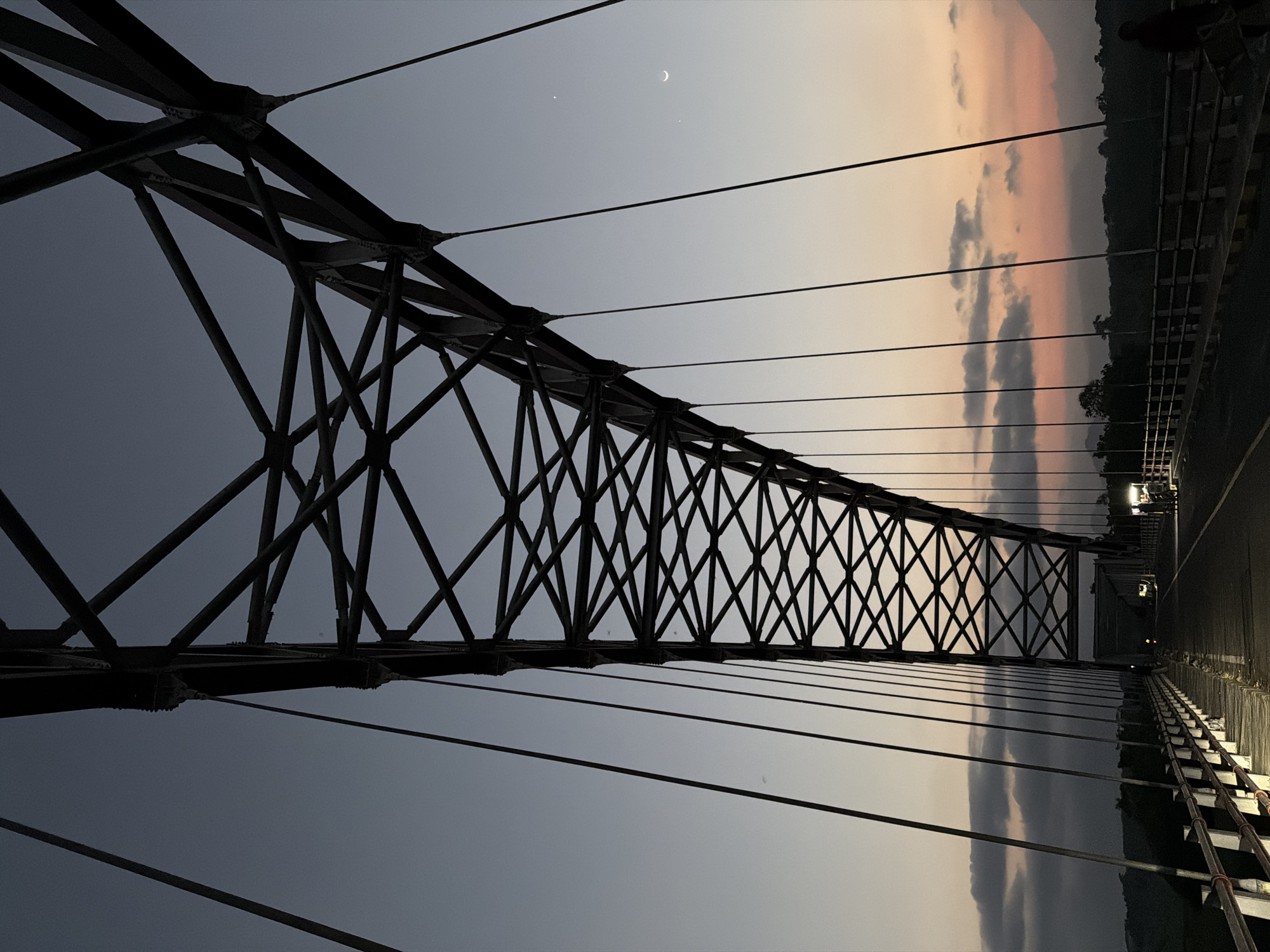 The image captures a suspension bridge at dusk in Selokanayar, Indonesia. The primary subjects are the intricate steel structure of the bridge and the vast, fading sky. The bridge's framework, made of dark metal beams and cables, dominates the foreground and midground, creating a dramatic geometric pattern against the horizon. Suspension cables stretch diagonally across the scene, anchoring the structure.
In the sky, the transition from day to night is evident. The sky is painted with soft hues of pale grey and a gentle peach, suggesting the last light of sunset. Wispy clouds are scattered across the upper portion of the sky, and a crescent moon is visible, accompanied by two faint stars. The landscape beyond the bridge is silhouetted against the sky, appearing as a dark, forested ridge. The roadway of the bridge is visible on the right, illuminated by some distant lights, indicating a road or walkway.
There is no discernible activity or event taking place; the image appears to be a static capture of the bridge and its surroundings during twilight. The mood is serene and peaceful, with the muted colors and the quiet grandeur of the bridge contributing to a sense of calm. The time of day is clearly evening, as indicated by the sunset colors and the visible celestial bodies. The location is identified as Selokanayar, Indonesia, though no specific landmarks are immediately recognizable beyond the bridge itself. No text is visible in the image.