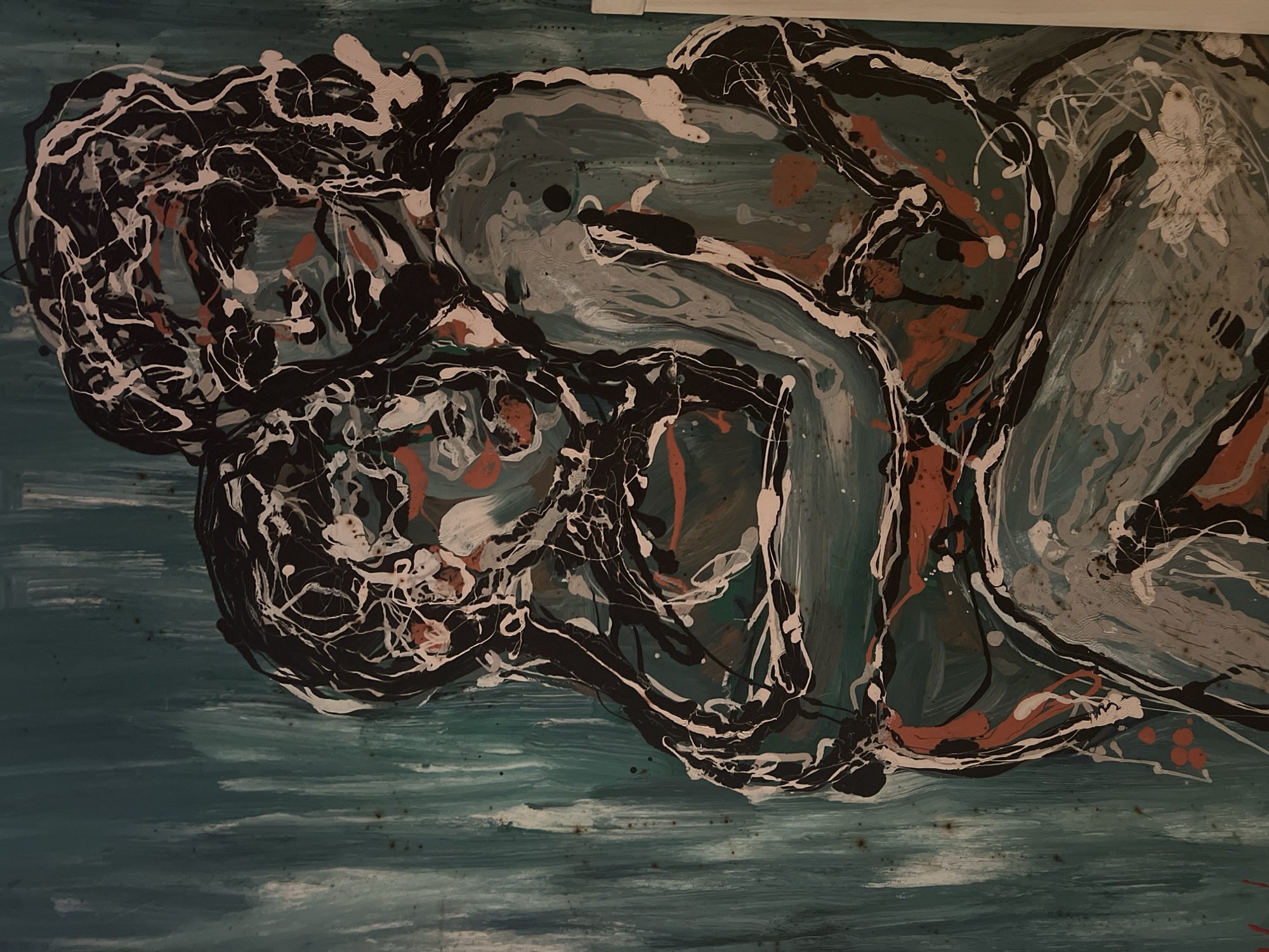 The image is a close-up of an abstract painting. The painting features two figures embracing, rendered in a chaotic, expressive style with bold brushstrokes and splatters of black, white, and orange paint over a teal background. The background itself is textured with vertical, sweeping strokes of teal and lighter greenish-blue. The figures are not clearly defined but suggest a human form through their intertwined limbs and heads. One figure appears to be cradling the other. The overall impression is one of intense emotion and connection. The location of this painting within Acquaviva, Italy, is not determinable from the image itself.
