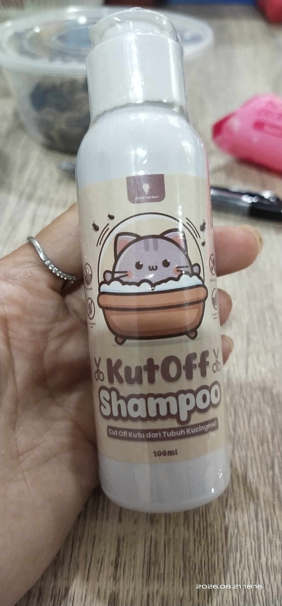 The image shows a bottle of "Kut Off Shampoo" held in a person's hand. The bottle has a cartoon image of a cat in a bathtub, surrounded by illustrations of fleas and scissors. The text on the bottle is in Indonesian and translates to "Cut Off Fleas from Your Cat's Body". The bottle is labeled as 100ml. The background appears to be a wooden surface, and a blurred container is visible behind the bottle. The timestamp at the bottom right of the image indicates the date and time are 2026.08.21 18:16. The location is identified as Matabu, Indonesia.