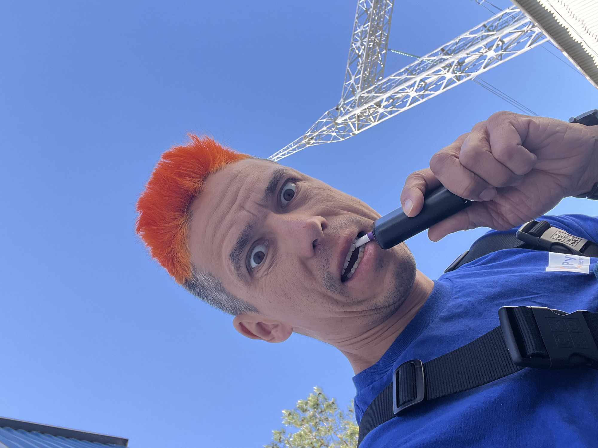 A low-angle, close-up shot depicts a man with orange hair, a blue t-shirt, and black straps across his chest holding a black object in his mouth. The man looks directly at the camera with a surprised expression. Above him, a clear blue sky is visible, along with a white metal structure resembling a power transmission tower. In the lower left corner, a section of a building roof is visible, and in the lower right corner, there is a tree with green leaves.
The location is identified as Chaikovsky, Russia.