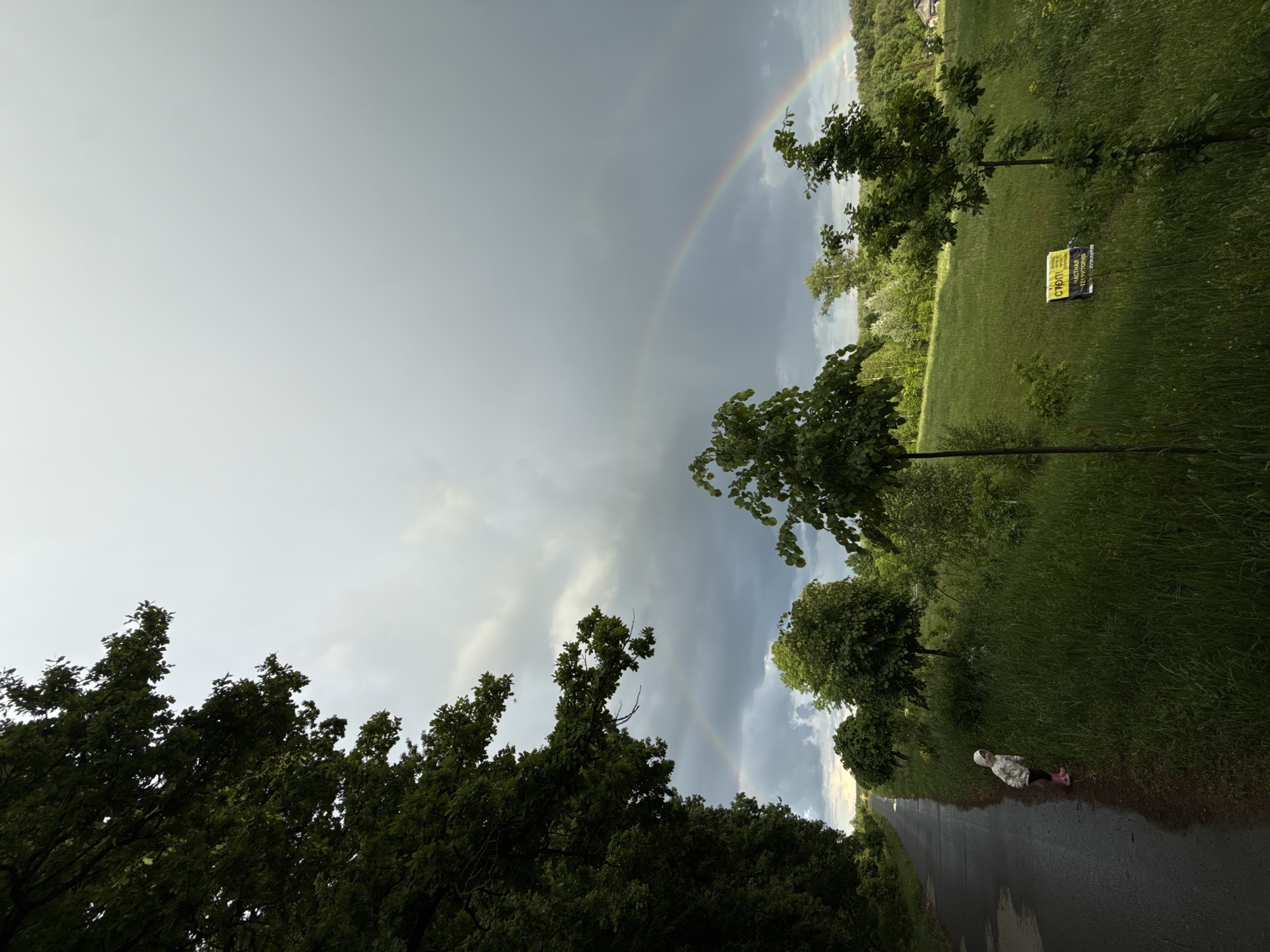 Captured in Sapronovo, Russia, the image presents a double rainbow arching across a stormy sky. The landscape below is characterized by lush green grass, various trees, and a sign placed on the ground. A wet road runs into the distance, alongside which a small child in a white and patterned coat and pink boots is walking.