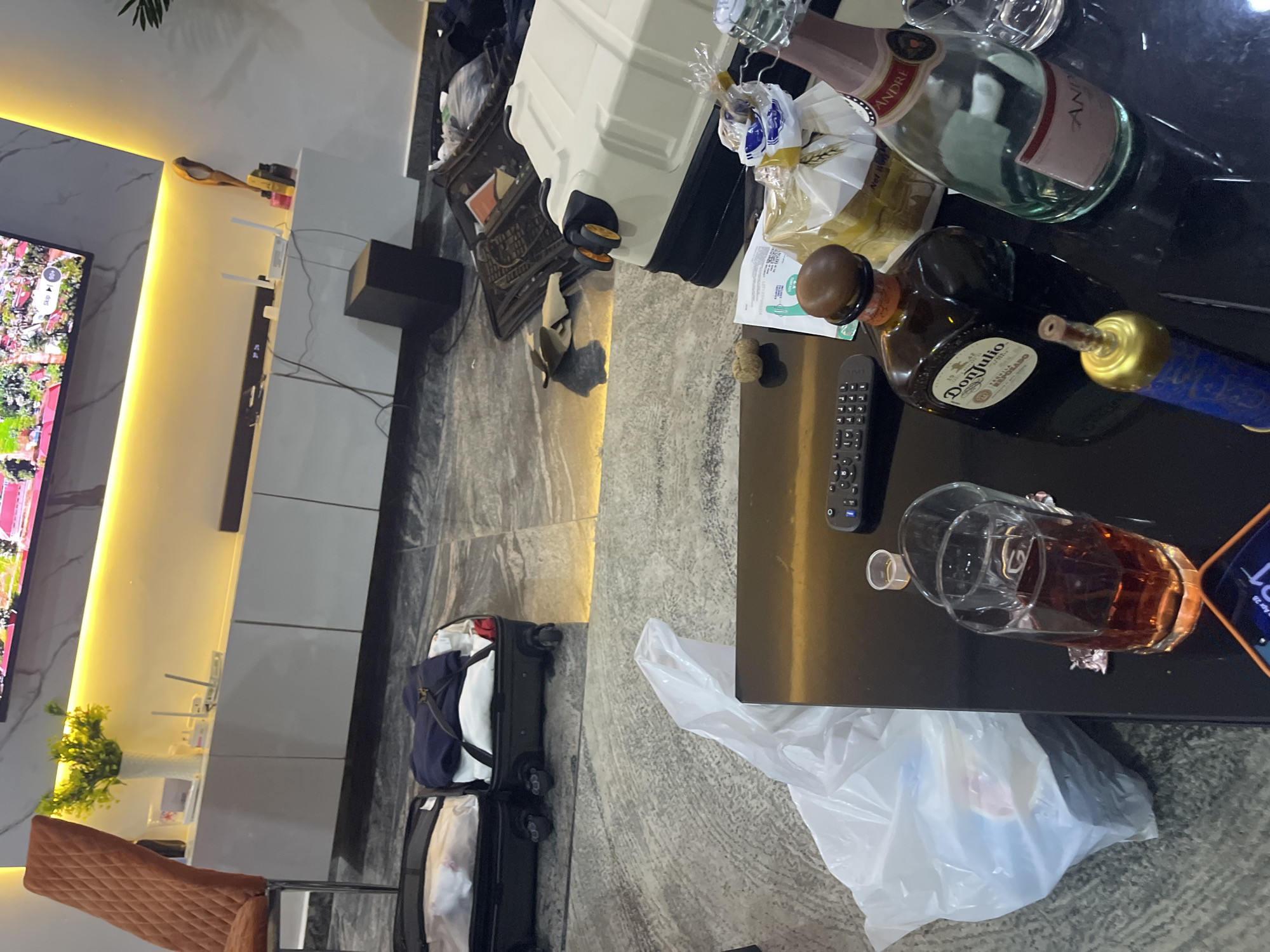 The image captures an indoor scene, likely a living room or hotel room, characterized by modern decor and a slightly disheveled state, suggesting recent activity or travel.
On the right side, a black, glossy coffee table holds several items. Prominently displayed are various alcoholic beverages: an empty green bottle of "André" sparkling wine, a brown bottle of "Don Julio" tequila, and a distinctive blue and gold bottle, possibly another spirit. A glass containing an amber liquid, presumably a drink, is also on the table, alongside a small clear plastic cup. A TV remote control and a partially visible mobile phone screen showing "Apr 28" and the time "1:31" are also present. A white plastic bag, possibly containing refuse, rests on the floor next to the table.
The background reveals a large flat-screen television displaying colorful content, possibly an outdoor scene with lush greenery. Below the TV is a sleek, white media console featuring an integrated yellow LED light strip that illuminates the area. On the console are a black speaker or subwoofer, two white Wi-Fi routers with visible antennae, and a decorative plant in a white vase. Wires are visible behind the console. The wall behind the console has a subtle marble-like pattern.
In the mid-ground, near the console, are several pieces of luggage, some open with clothes visible inside, indicating packing or unpacking. An orange-brown upholstered chair with a quilted pattern is visible on the far left. The floor throughout the room is a mix of grey and white patterned tiles or polished concrete. No people are present in the image. The arrangement of items suggests a casual atmosphere, potentially after a social gathering or during a travel transition.