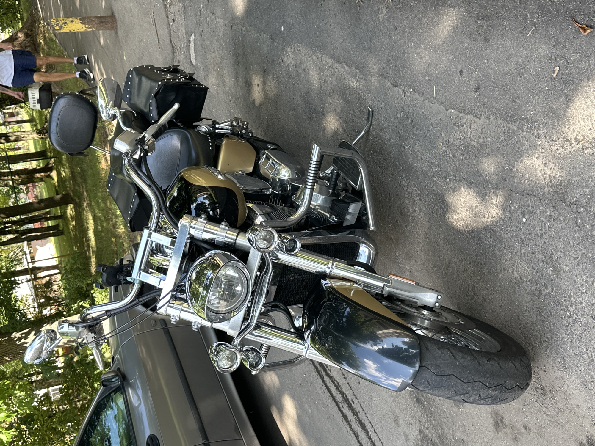 The image shows a motorcycle parked on an asphalt surface, likely a street or sidewalk in Minsk, Belarus. The motorcycle is a cruiser-style bike, predominantly black and gold, with chrome accents. It has a large headlight, dual auxiliary lights, a black leather seat with studs, and two studded leather saddlebags. A silver car is parked to the left of the motorcycle, and in the background, there are trees, grass, and a building. A person is walking in the background, carrying a pet carrier. The lighting suggests it is daytime, with sunlight filtering through the trees, casting shadows on the ground.