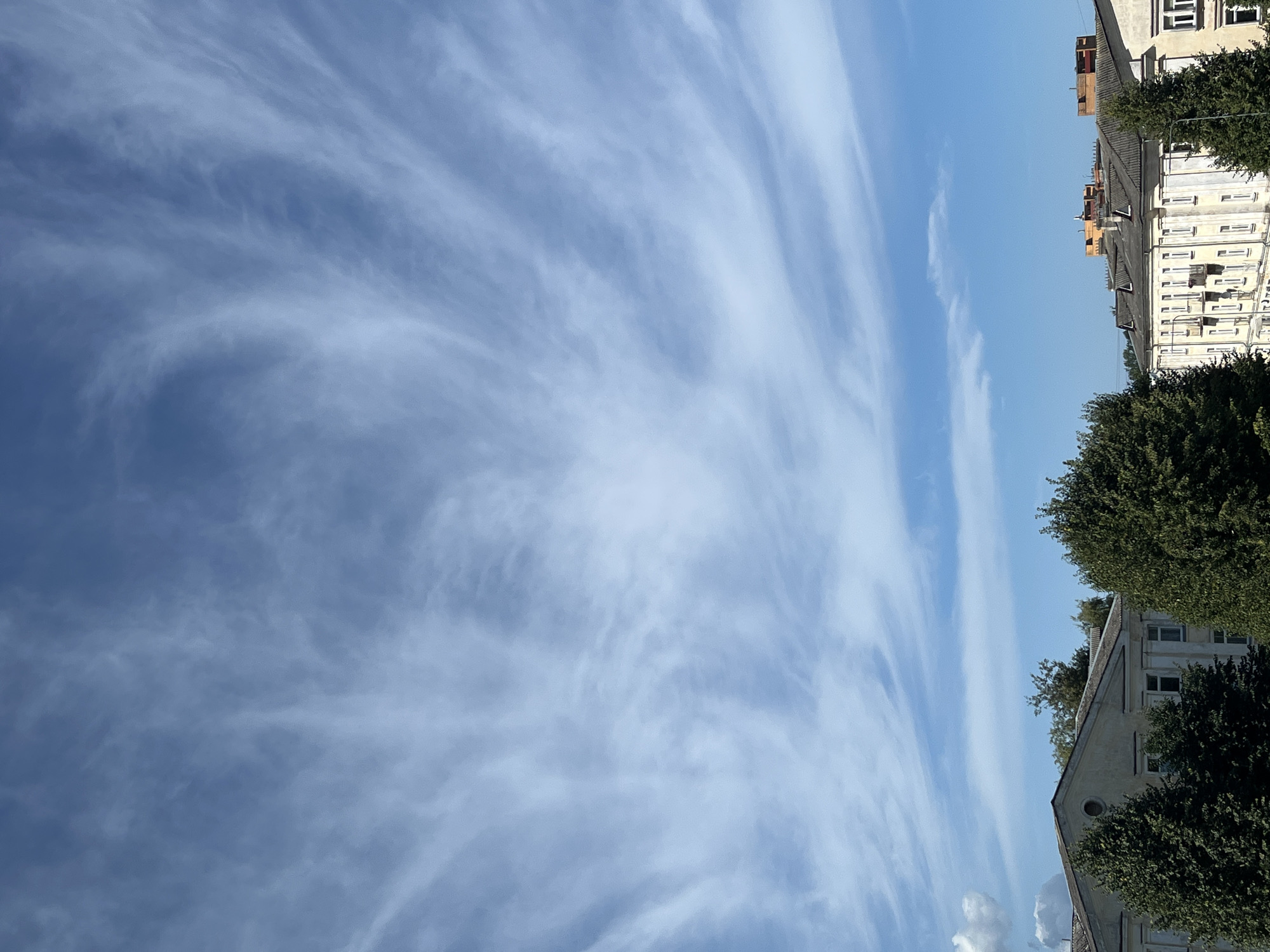 The image captures a view of buildings and trees under a sky with wispy clouds. The sky is a bright blue with thin, streaky clouds that create a sense of movement. In the foreground, the tops of several green trees are visible. Behind the trees are the roofs and upper stories of buildings. The architecture appears to be residential or commercial, with multiple windows and some balconies visible. The roofs are dark and sloped. The scene suggests an urban environment in Kohtla-Järve, Estonia, during daylight. There are no people or animals visible in the image. The overall impression is one of a calm, clear day.