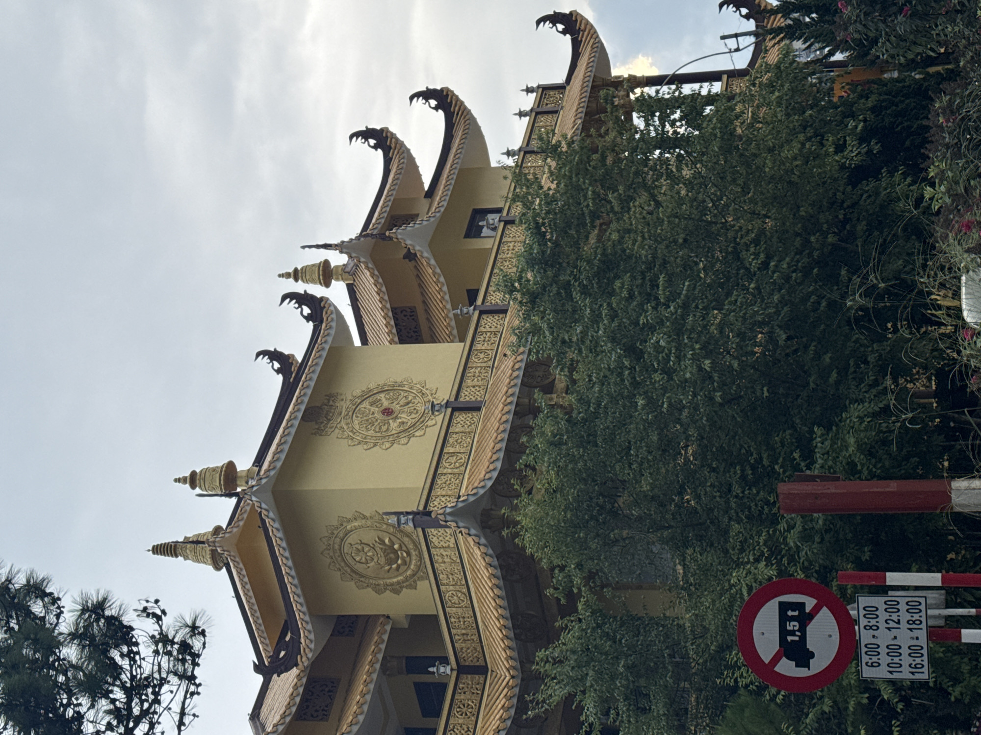This is a low-angle, portrait-oriented photograph of a building with distinctive East Asian architectural elements, possibly a temple or pagoda, located in Da Lat, Vietnam. The building features ornate, multi-tiered roofs with curved eaves that are decorated with what appear to be dragon-like sculptures or finials. The facade is painted a pale yellow and adorned with intricate gold patterns, including circular motifs with lotus-like designs and decorative borders.
In the foreground and to the right of the building, there are lush green trees, suggesting an outdoor setting. Partially visible on the right edge of the frame are traffic signs. One sign is circular with a red border and a diagonal line through it, indicating a prohibition. Inside the circle, there's an image of a truck and the text "1,5 t," likely signifying a weight limit. Below this is a rectangular sign with text indicating restricted hours for vehicles: "6:00-8:00", "10:00-12:00", and "16:00-18:00". The sky is overcast, indicating a potentially cloudy or diffused daylight. The image's perspective and the architecture suggest a cultural or religious site.