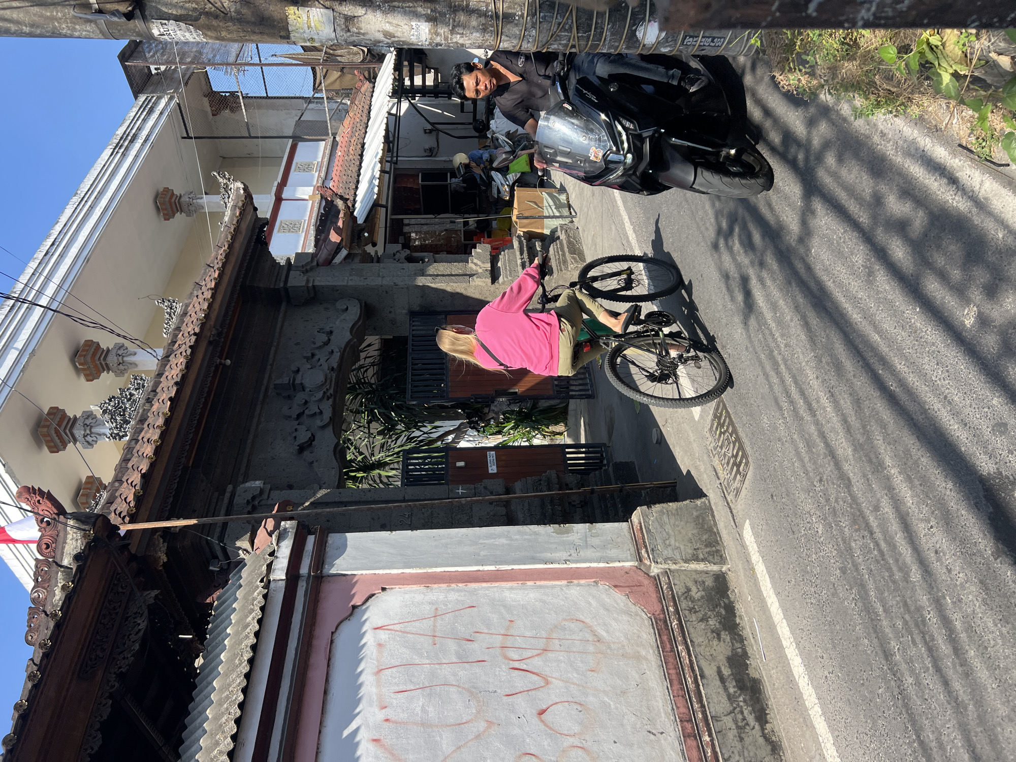 The image depicts a street scene in Kuta, Indonesia, under a clear blue sky. A woman with blonde hair, wearing a pink sweatshirt and khaki shorts, is cycling a black bicycle down the road. She has a black bag slung over her shoulder and is wearing flip-flops. To her right, a man wearing a black shirt and jeans is riding a black motorcycle. The motorcycle has a large windshield and is positioned slightly ahead of the cyclist. The street is paved with asphalt and marked with a white line.
On the left side of the image, there is a wall with a pink border and white paint, bearing red graffiti that appears to read "KUTA BOYS". Behind the wall, an ornate stone gateway with a tiled roof and intricate carvings is visible. A white and red Indonesian flag is flying from the rooftop of a building in the background. The background also shows more buildings with tiled roofs and balconies, some with exposed brickwork and metal railings. Green foliage is visible peeking from behind the gateway. On the right side of the street, a rough-textured pole, possibly a utility pole, stands tall, with a weathered sign attached to it. A patch of green vegetation with some dry leaves is growing at the base of the pole. The sunlight casts distinct shadows across the road from the overhead structures and the people.